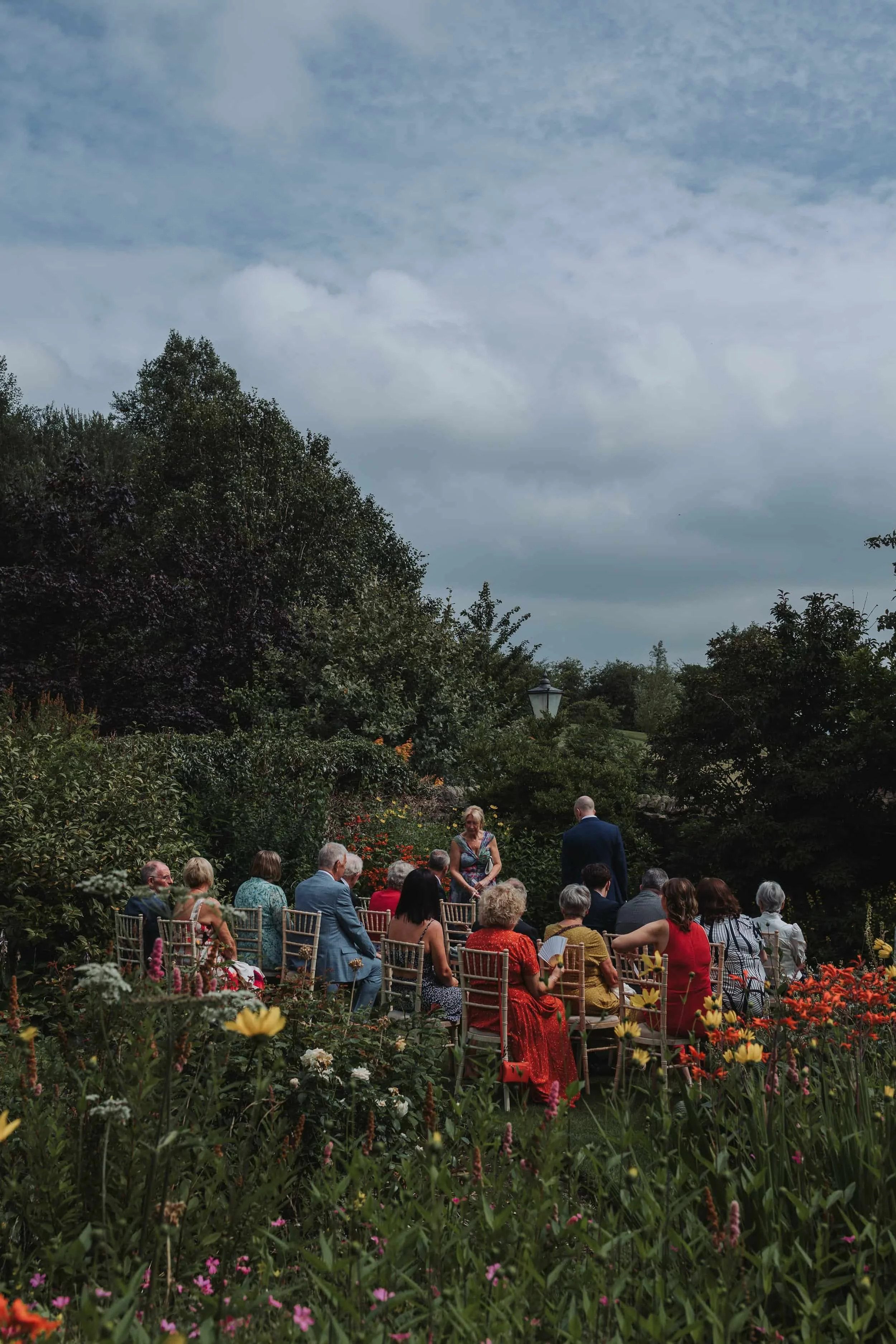 People attending an outdoor wedding ceremony in a lush garden with colorful flowers and trees under a partly cloudy sky.