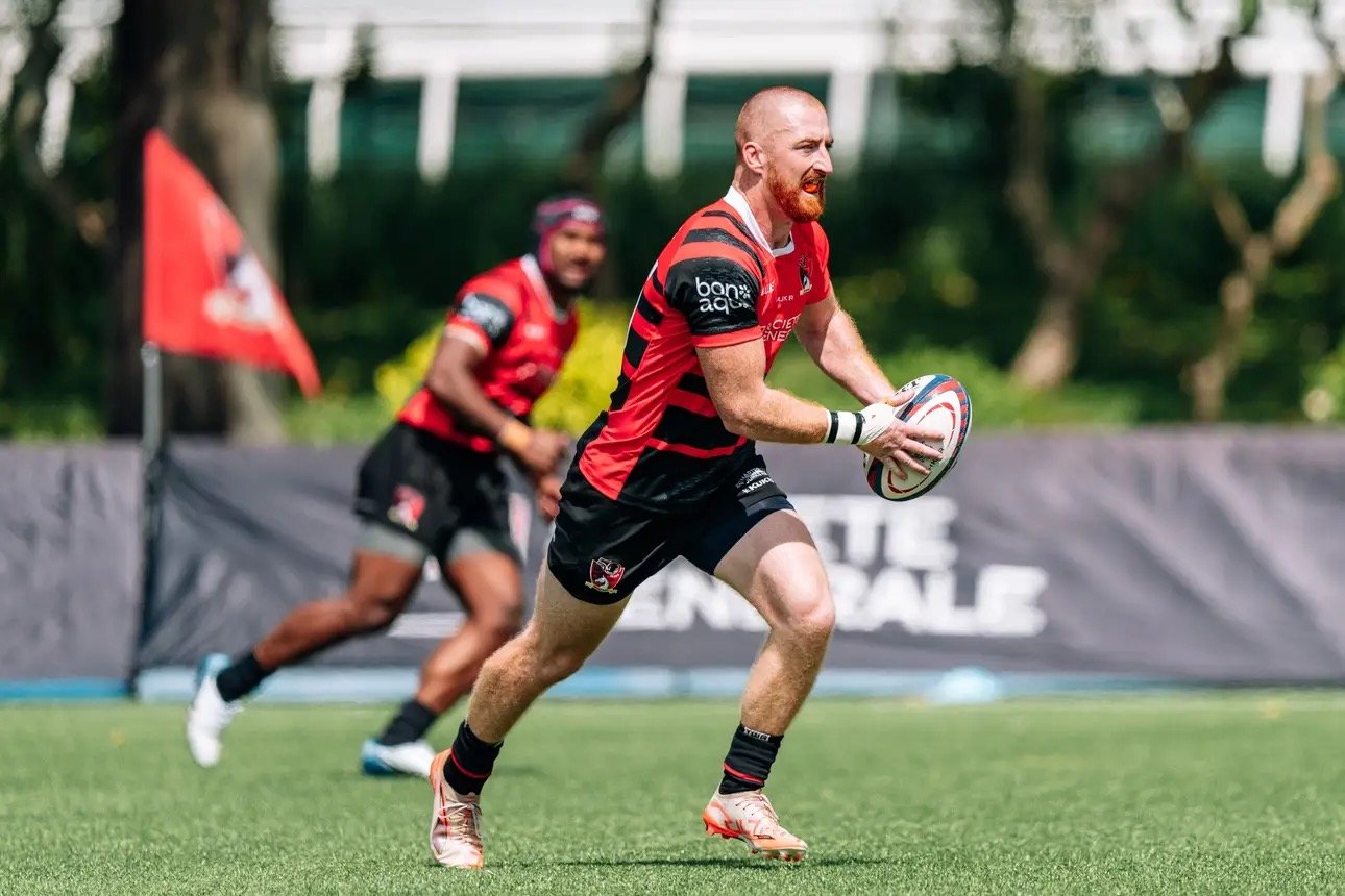 Rugby players in red and black uniforms during a game. One player is running with a rugby ball, while others are behind him.