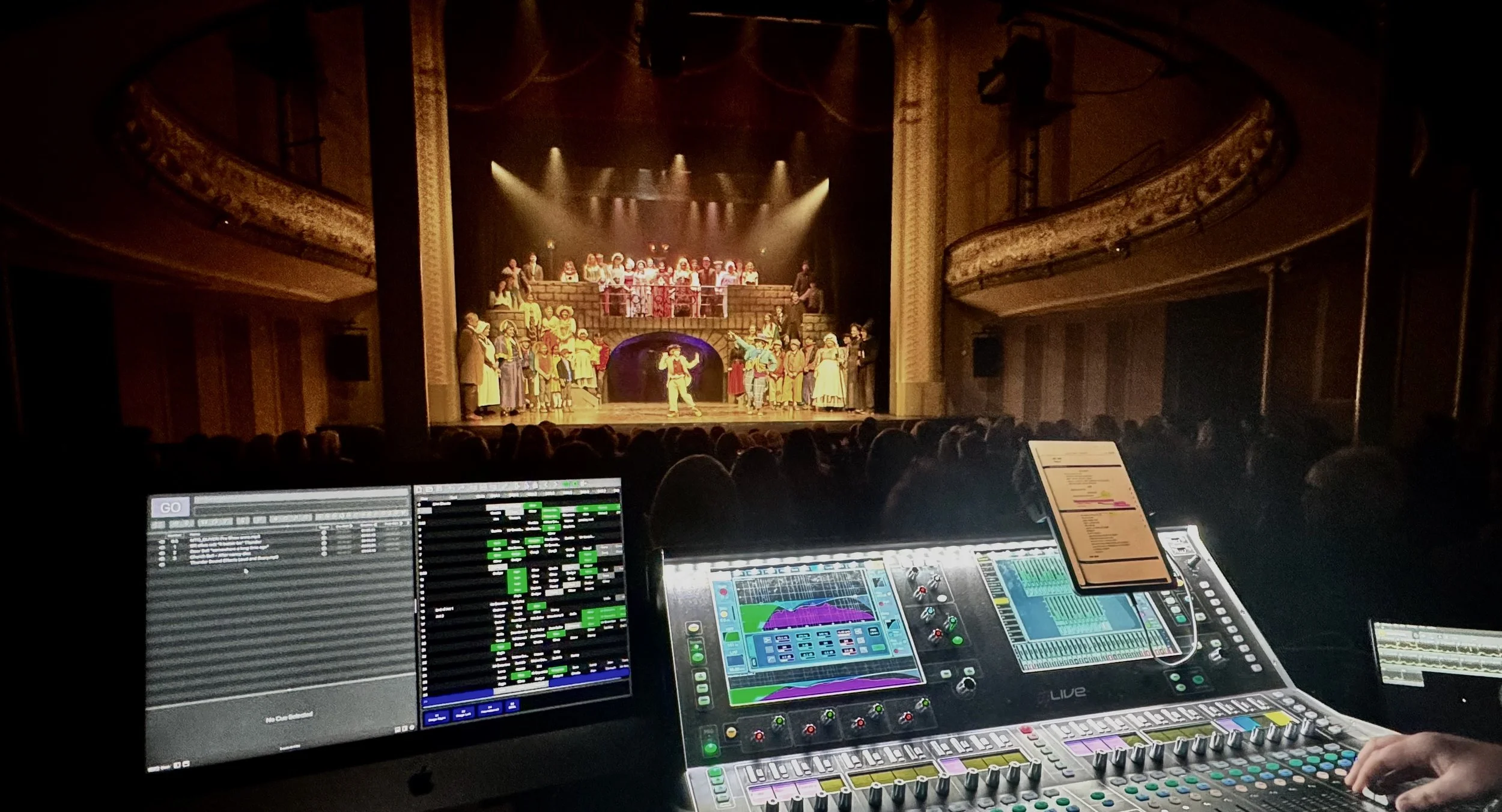 View of a Oamaru Opera House stage with a performance in progress, seen from the Sound desk, with an Allen & Heath Dlive in view.
