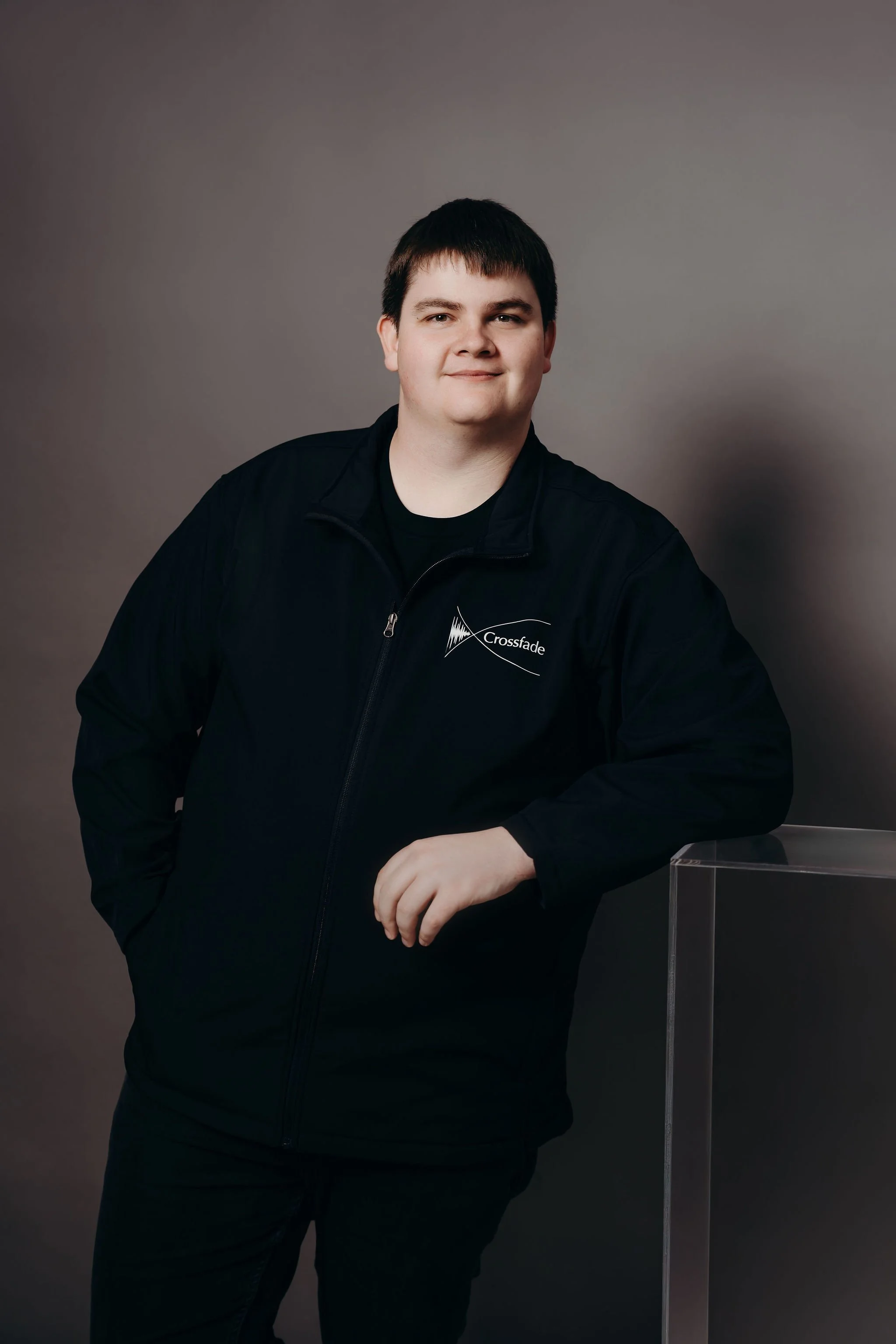 Tom Acklin from Dunedin with dark hair wearing a black jacket with a logo that reads 'Crossfade,' standing against a plain gray background, leaning on a clear acrylic podium.