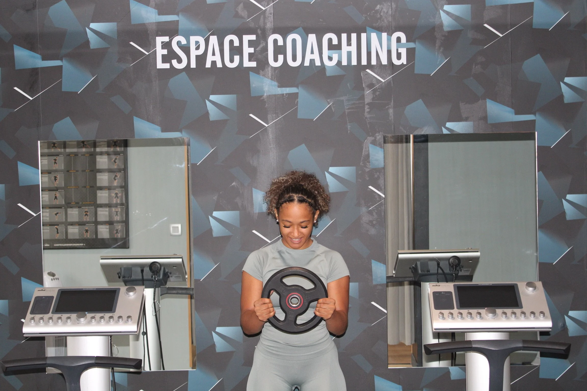 Une femme souriante en tenue de sport tient un disque de fitness devant un mur avec l'inscription 'ESPACE COACHING' dans une salle de sport. Il y a deux stations d'entraînement avec des équipements électroniques sur chaque côté.