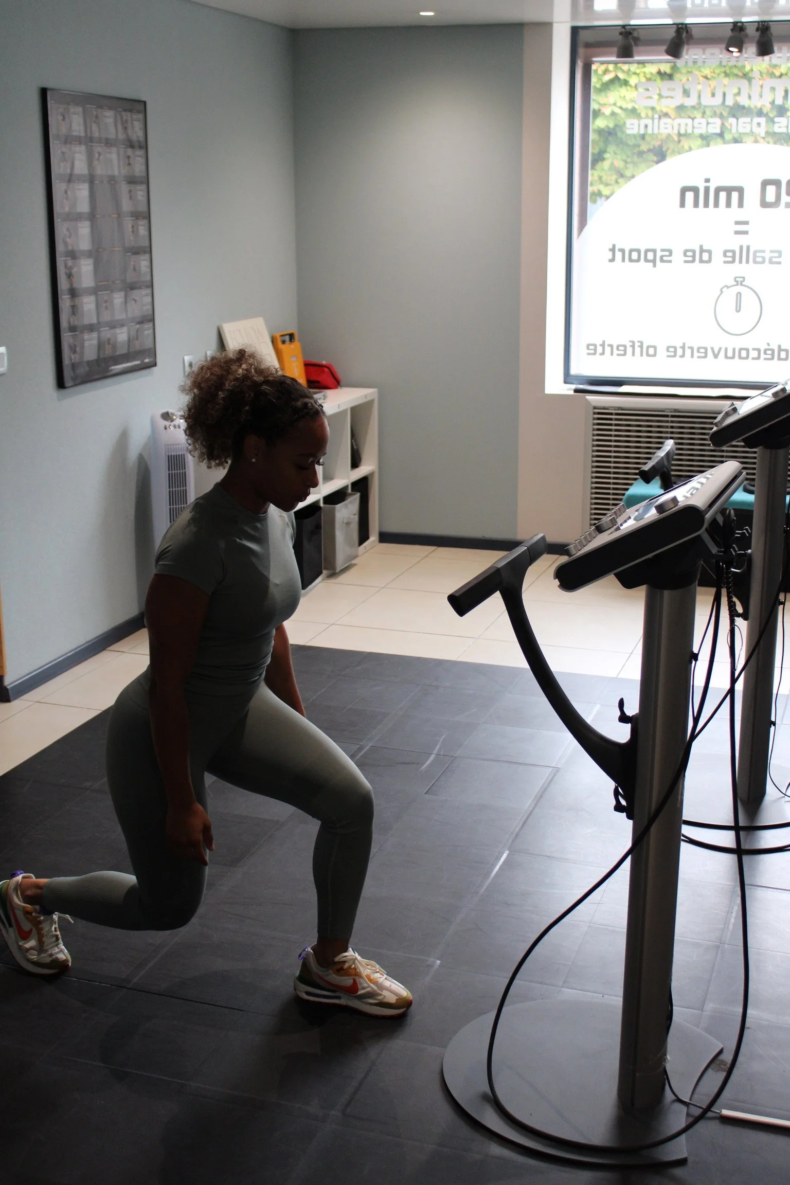 Une femme effectuant un exercice de fente dans une salle de sport près d'un tapis de course en tenue EMS SOSTIM