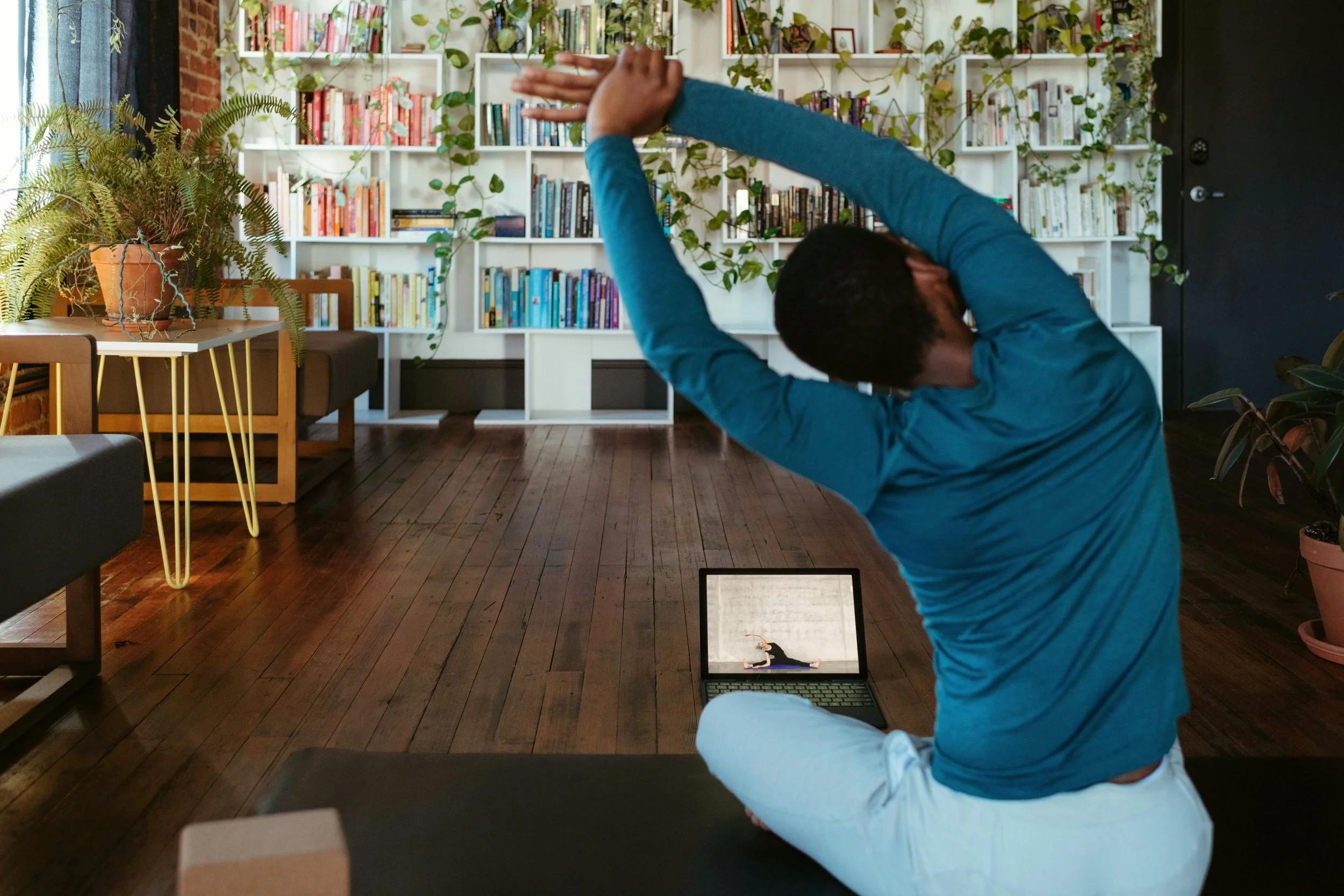 Une personne pratique le yoga assise sur un tapis dans un intérieur avec étagères remplies de livres.