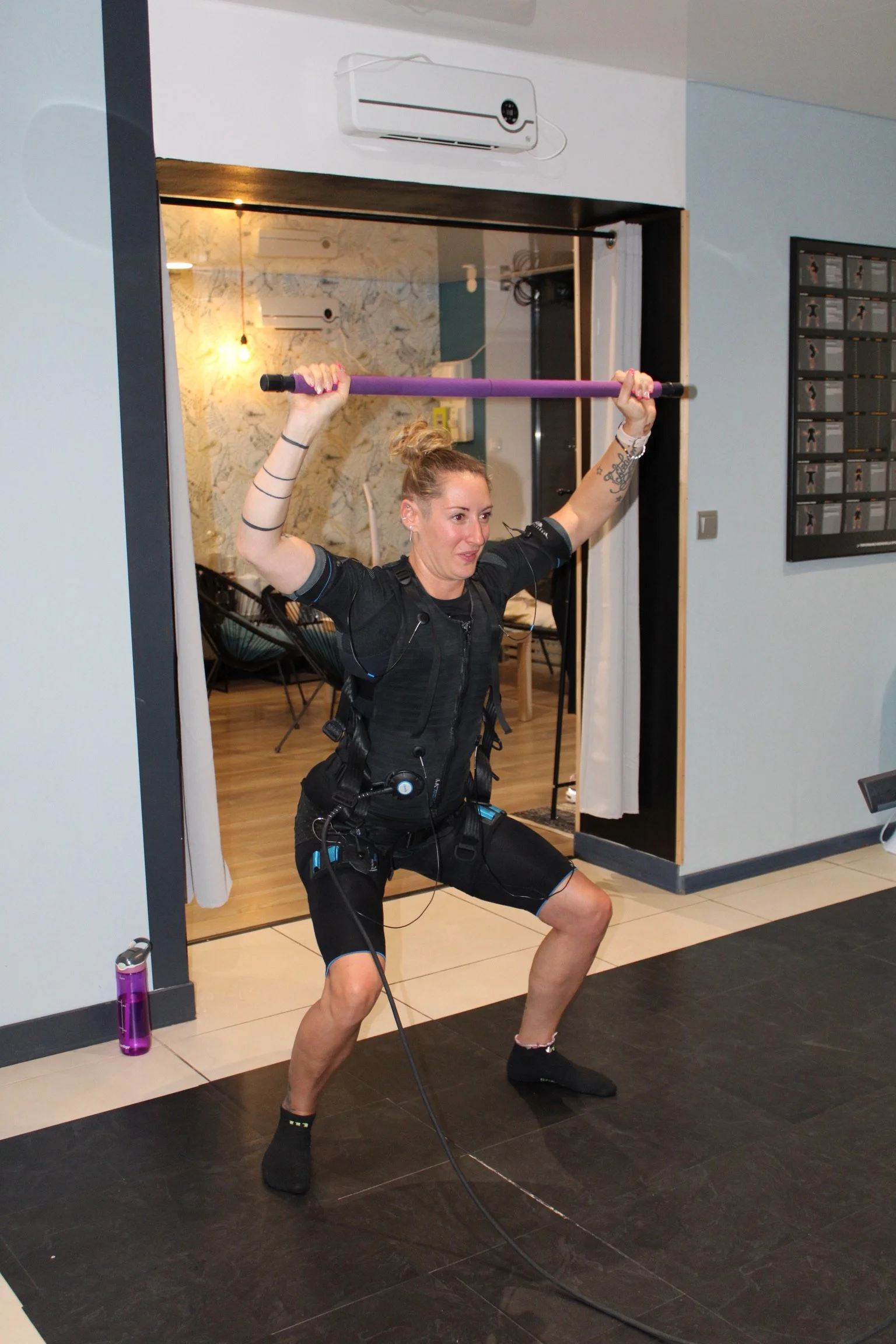 Femme en tenue d'EMS SOSTIM, faisant de l'exercice de squat avec un bâton dans un espace intérieur