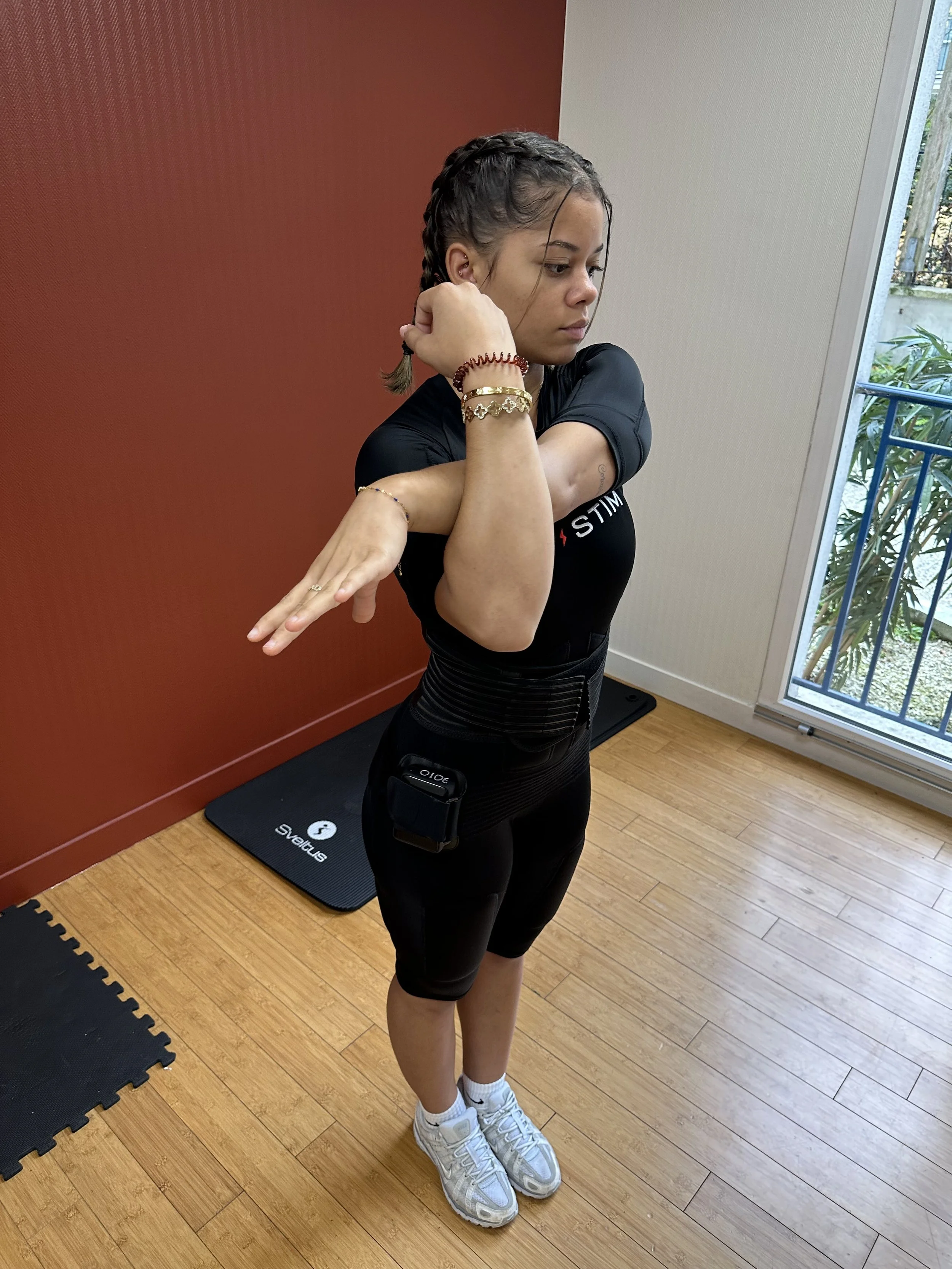 Une jeune femme en tenue de sport, portant des bracelets, en train de faire un exercice d'étirement ou de rotation du bras dans une pièce avec un sol en bois et une fenêtre.