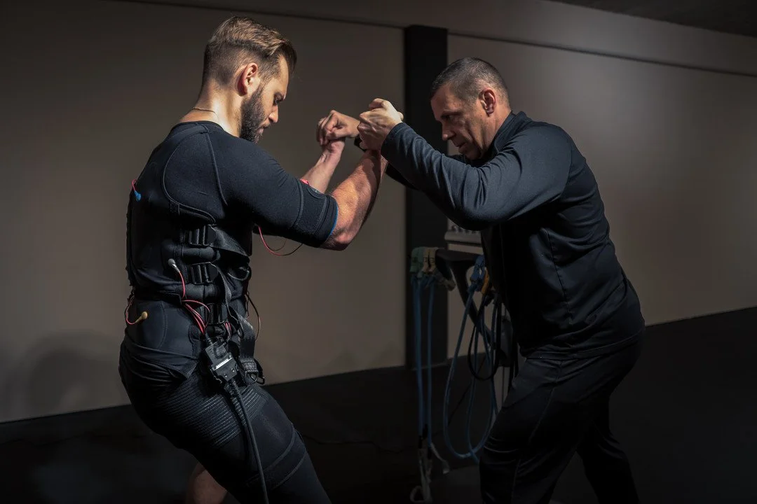 Deux hommes s'entraînent en combat simulé dans une salle de sport, l'un portant une tenue EMS SOSTIM