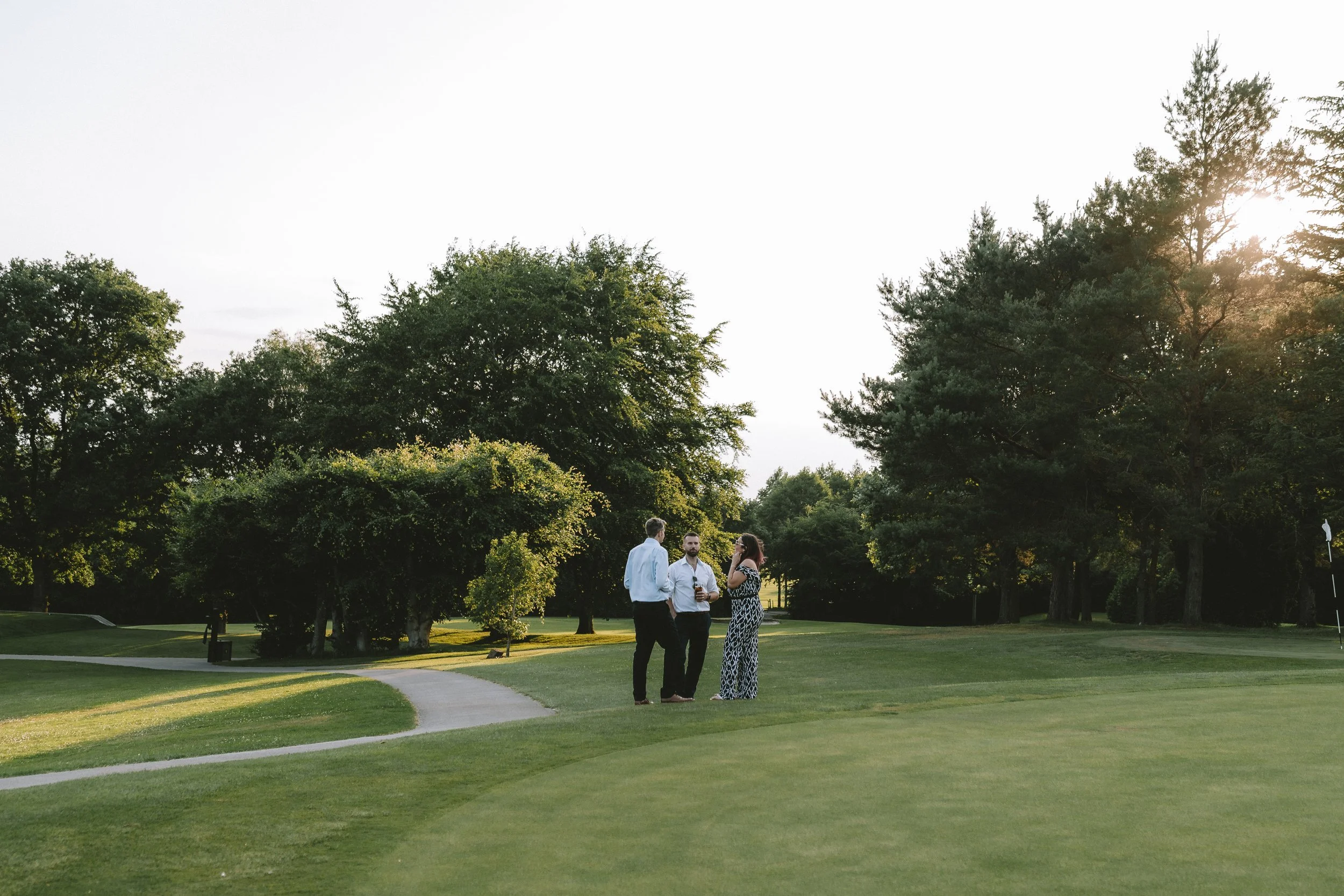 Four people standing on a well-maintained grassy park with tall trees in the background. The sun is setting, casting a warm glow. They are engaged in conversation, dressed in semi-formal attire.