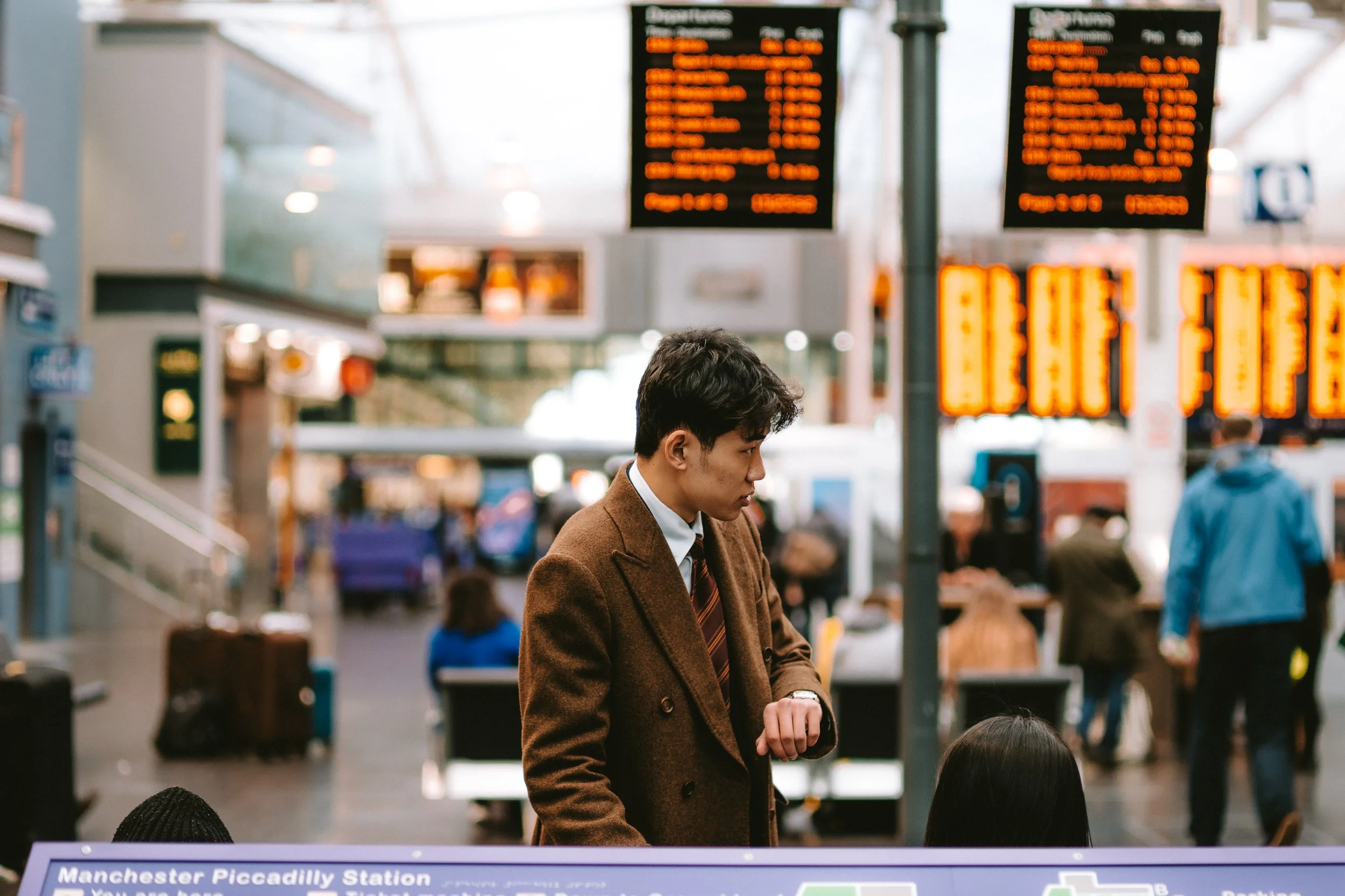 A young man in a brown blazer and striped tie looking at his watch at a busy train station with illuminated departure boards overhead.