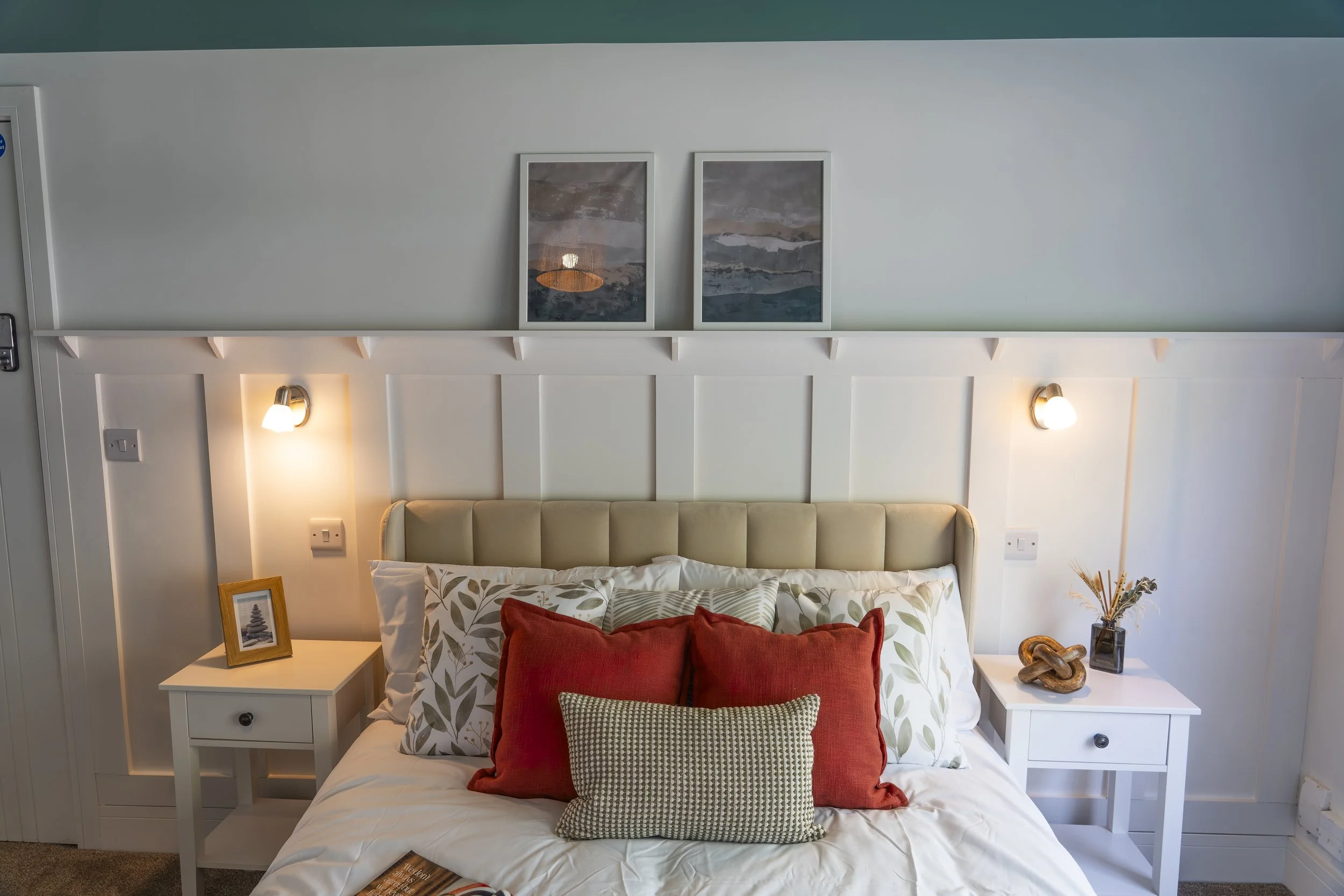 A cozy bedroom with a white bed, decorative pillows, white nightstands, framed artwork, and wall-mounted lamps.