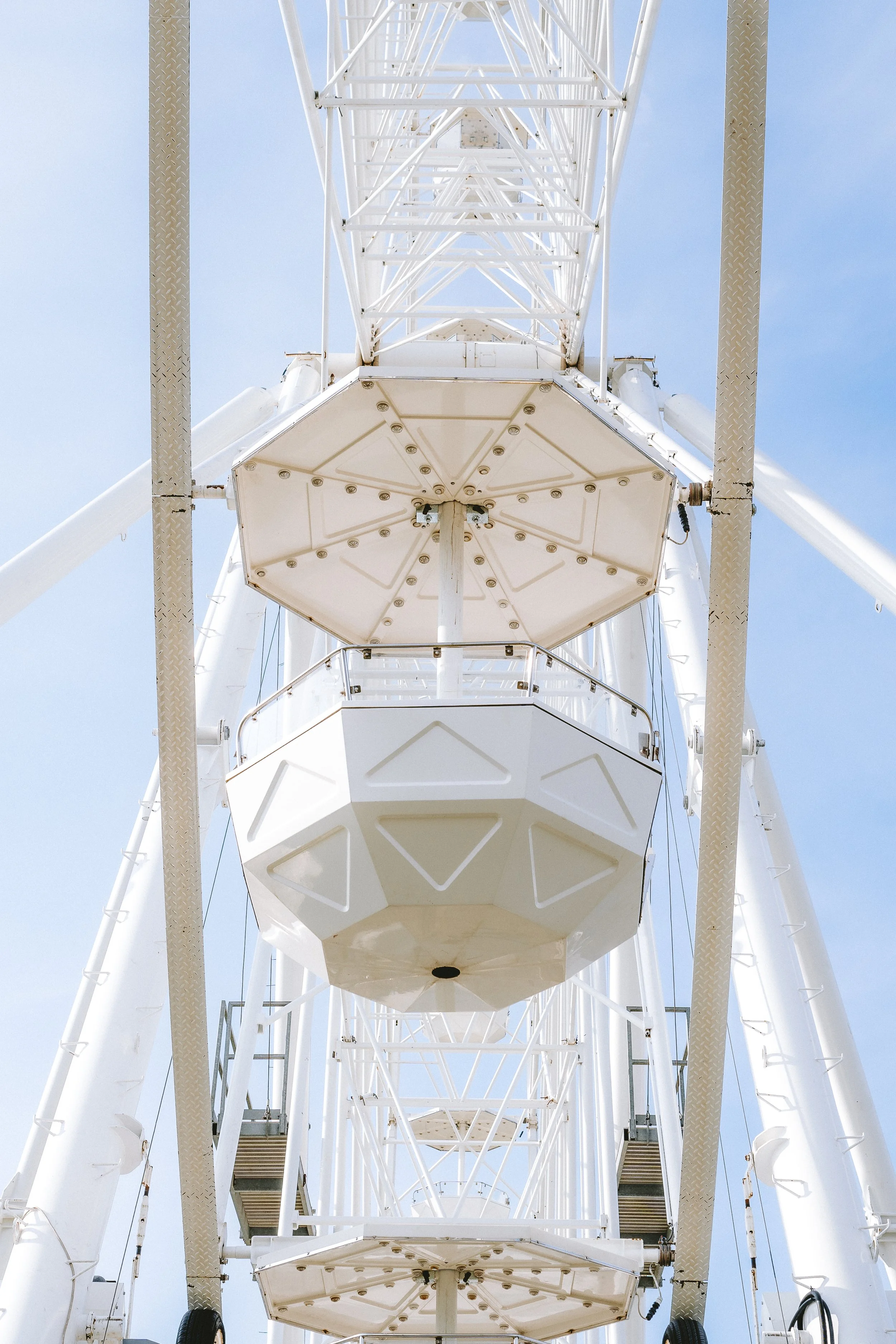 A view from below of a large white Ferris wheel with multiple passenger cabins visible, set against a clear blue sky.