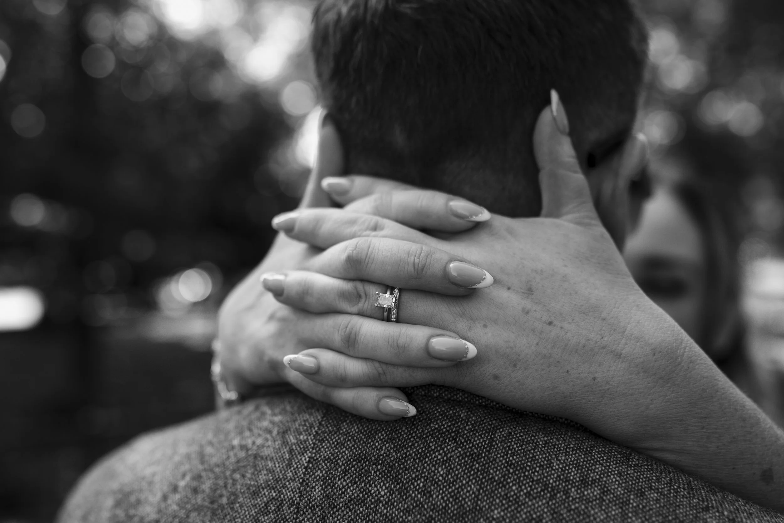 Close-up black and white photograph of a couple embracing, with the woman's hand and engagement ring visible, resting on the man's shoulder.