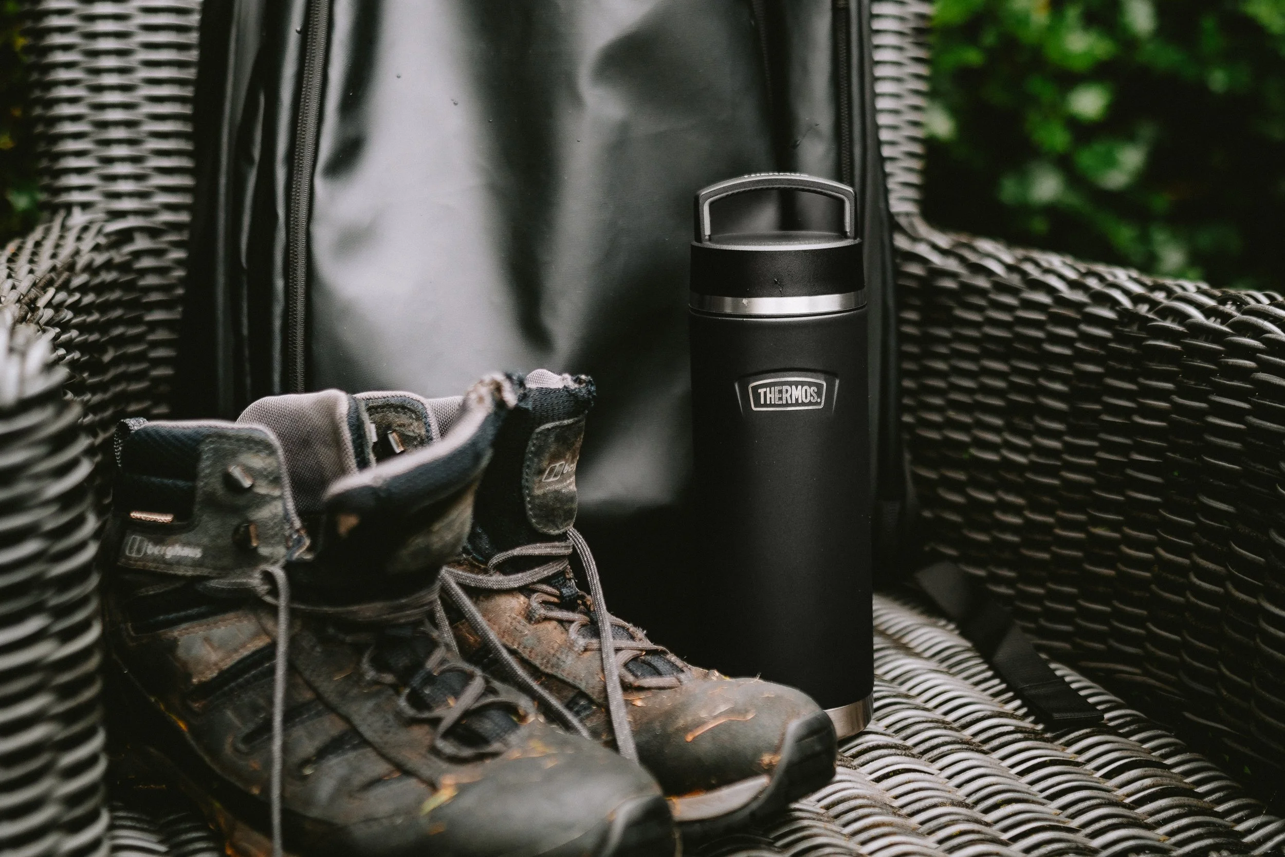 A pair of hiking boots, a black backpack, a Thermos flask, and a pair of outdoor gloves inside a woven basket with green foliage in the background.
