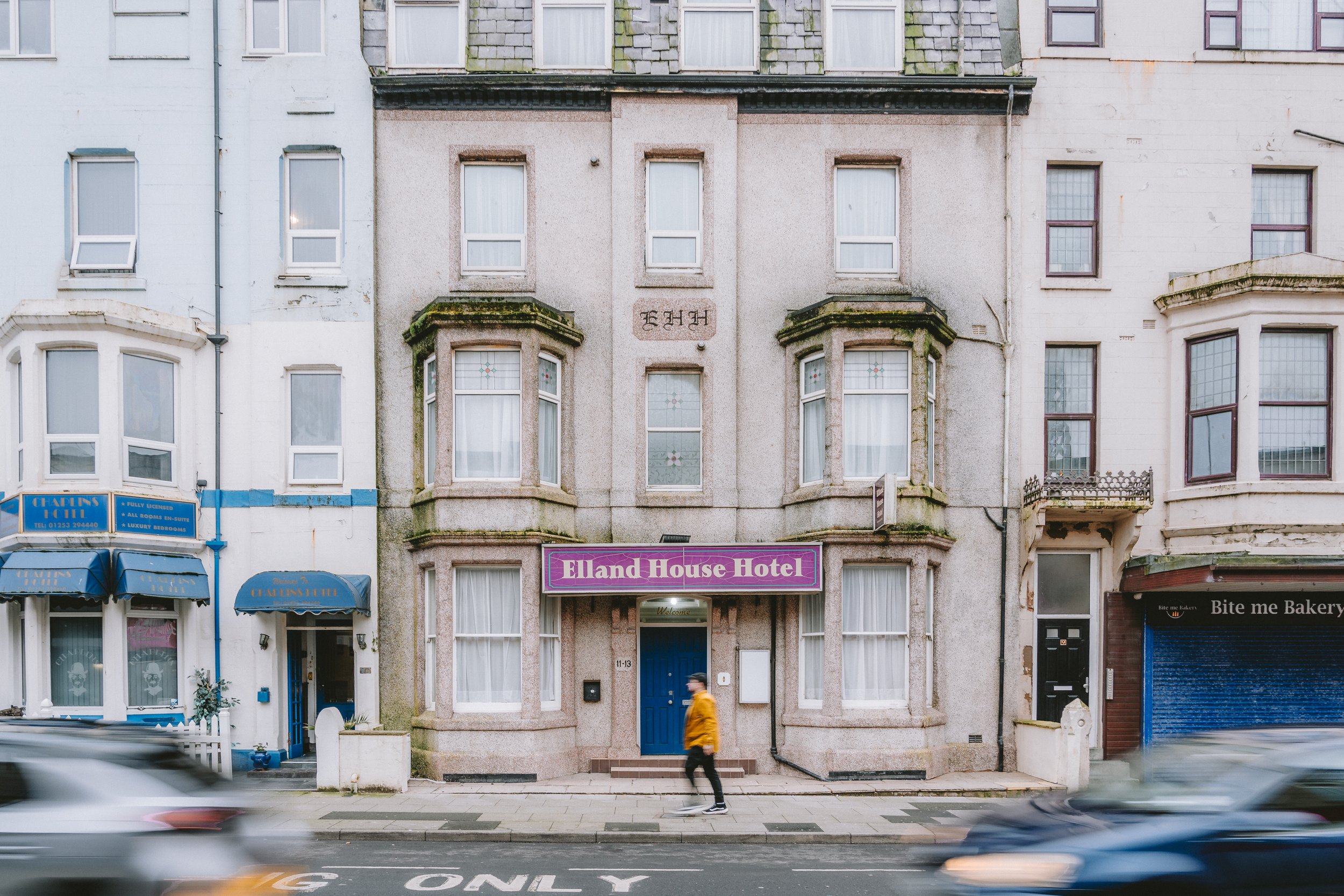 A multi-story building with a brick and concrete facade houses a hotel named Elland House Hotel. The hotel entrance has a blue door, a purple sign, and large windows with white curtains. The building is flanked by neighboring buildings, and shops are