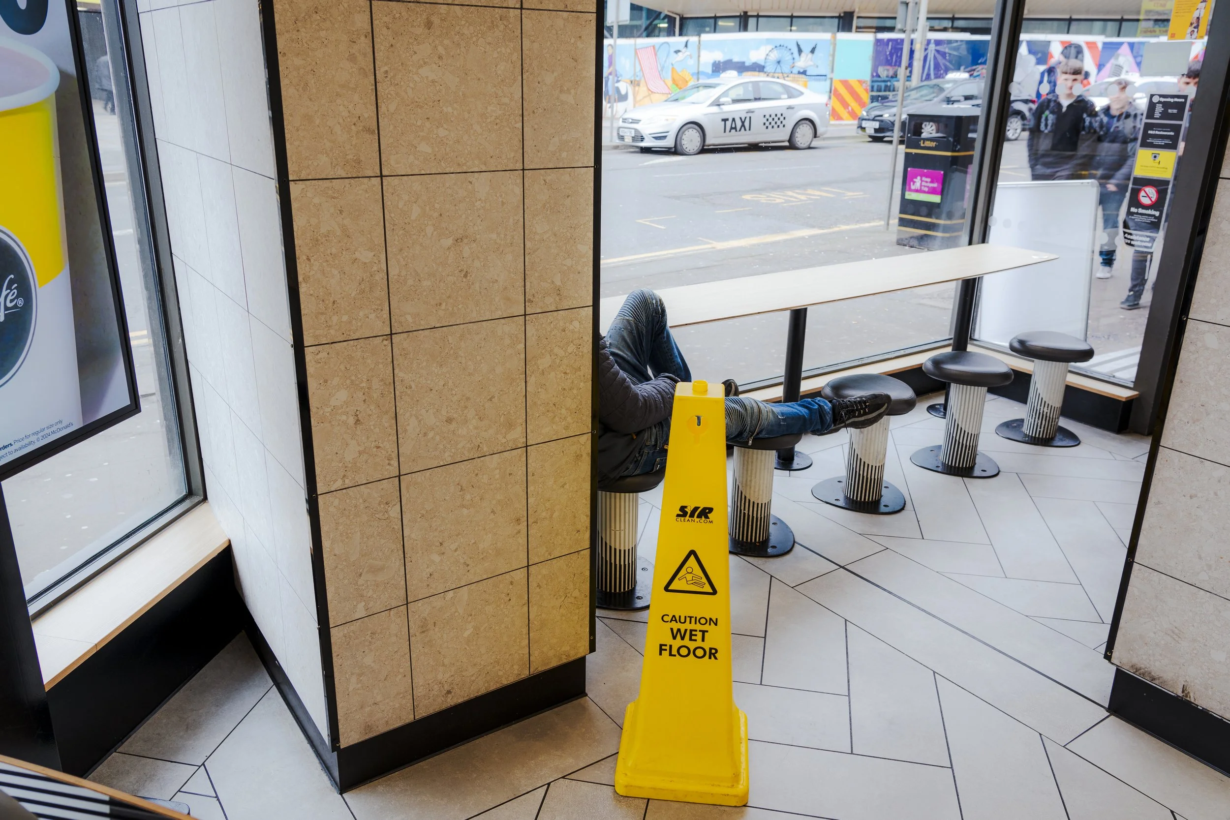 View from inside a fast food restaurant or cafe showing a wet floor caution sign and seating area. Outside, a taxi and a few pedestrians are visible through the glass windows.