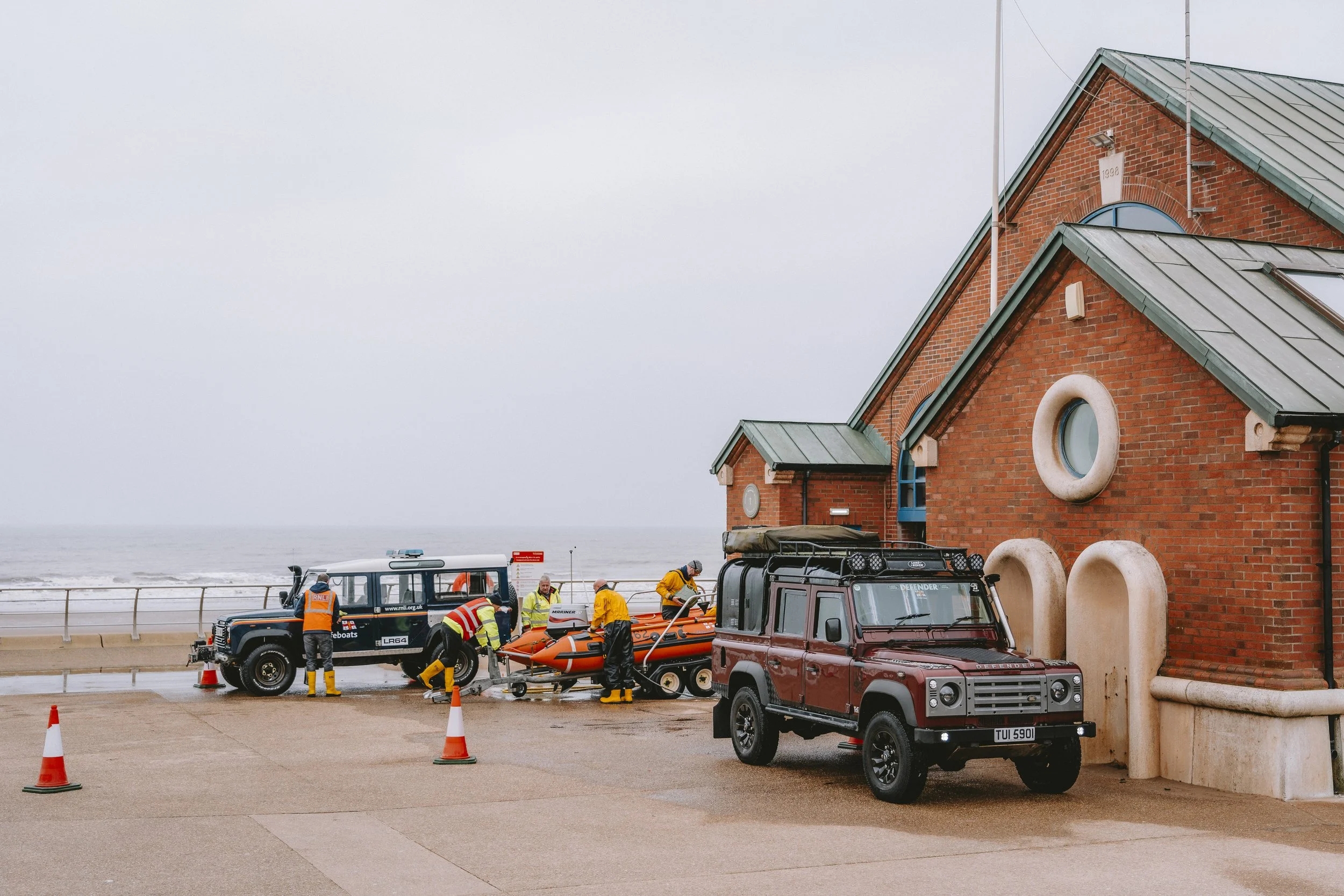 Lifeboat crew members preparing emergency response equipment near a brick building by the ocean, with a rescue boat and vehicles parked nearby, under an overcast sky.