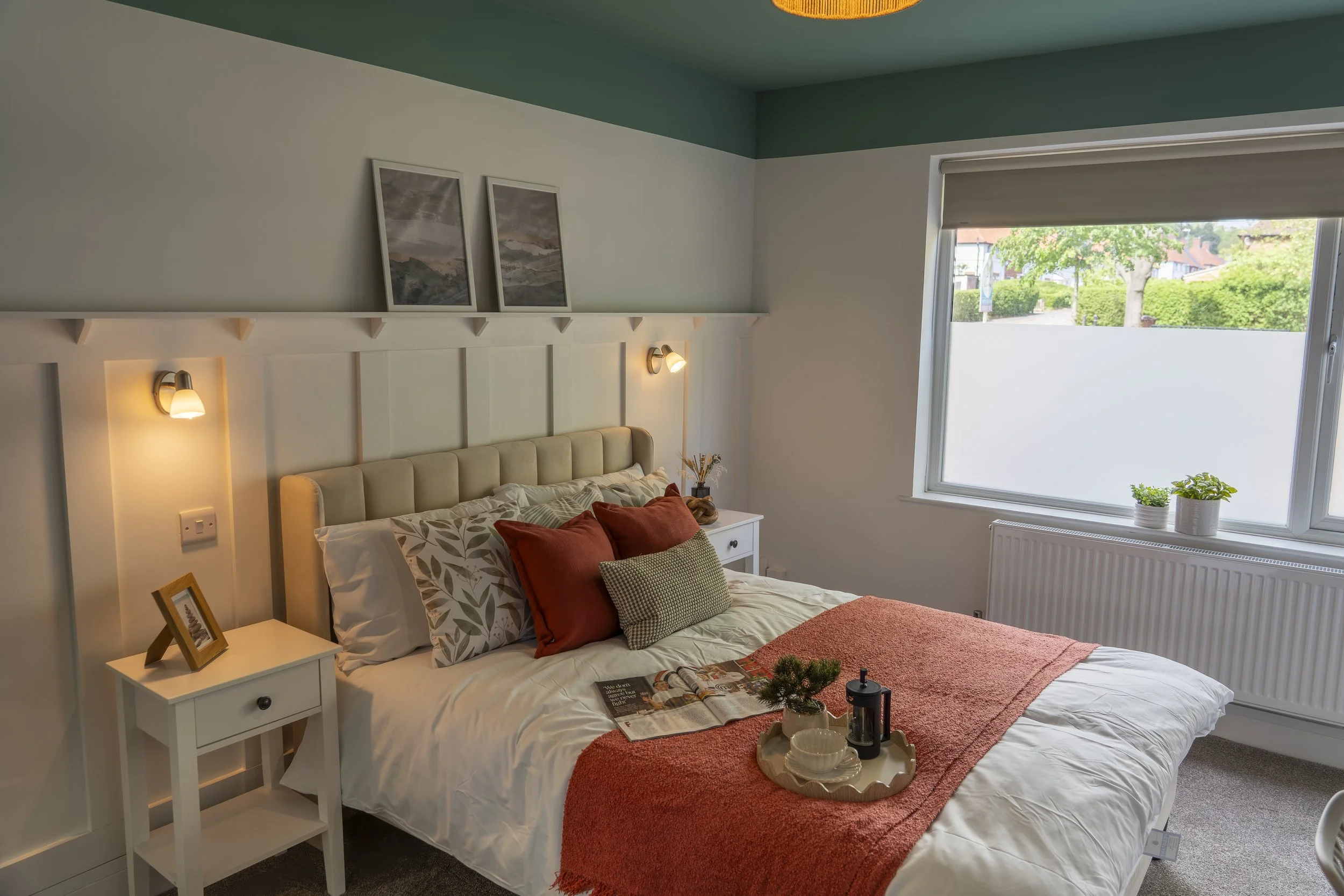 A cozy bedroom with a bed featuring a beige upholstered headboard, decorated with patterned and plain pillows, a coral-colored blanket, and a tray with cups and a small plant on it. There are two white nightstands, each with a framed photo and a smal