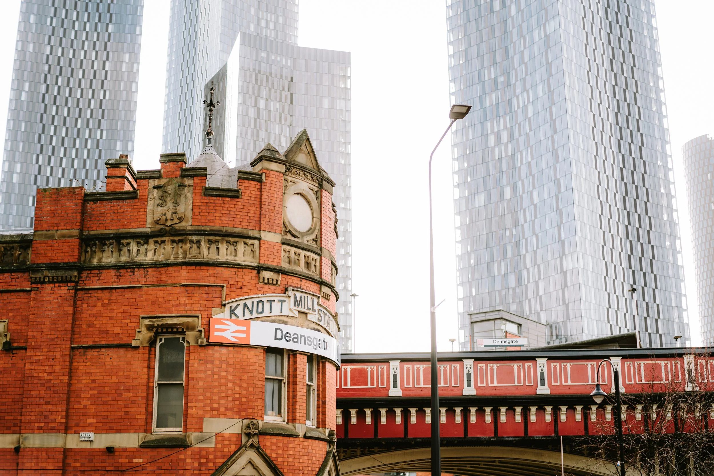 A historic red brick building with ornate architectural details and a cross on top, surrounded by modern glass skyscrapers in an urban setting, with street signs for Knott Street, Deansgate, and a subway sign.