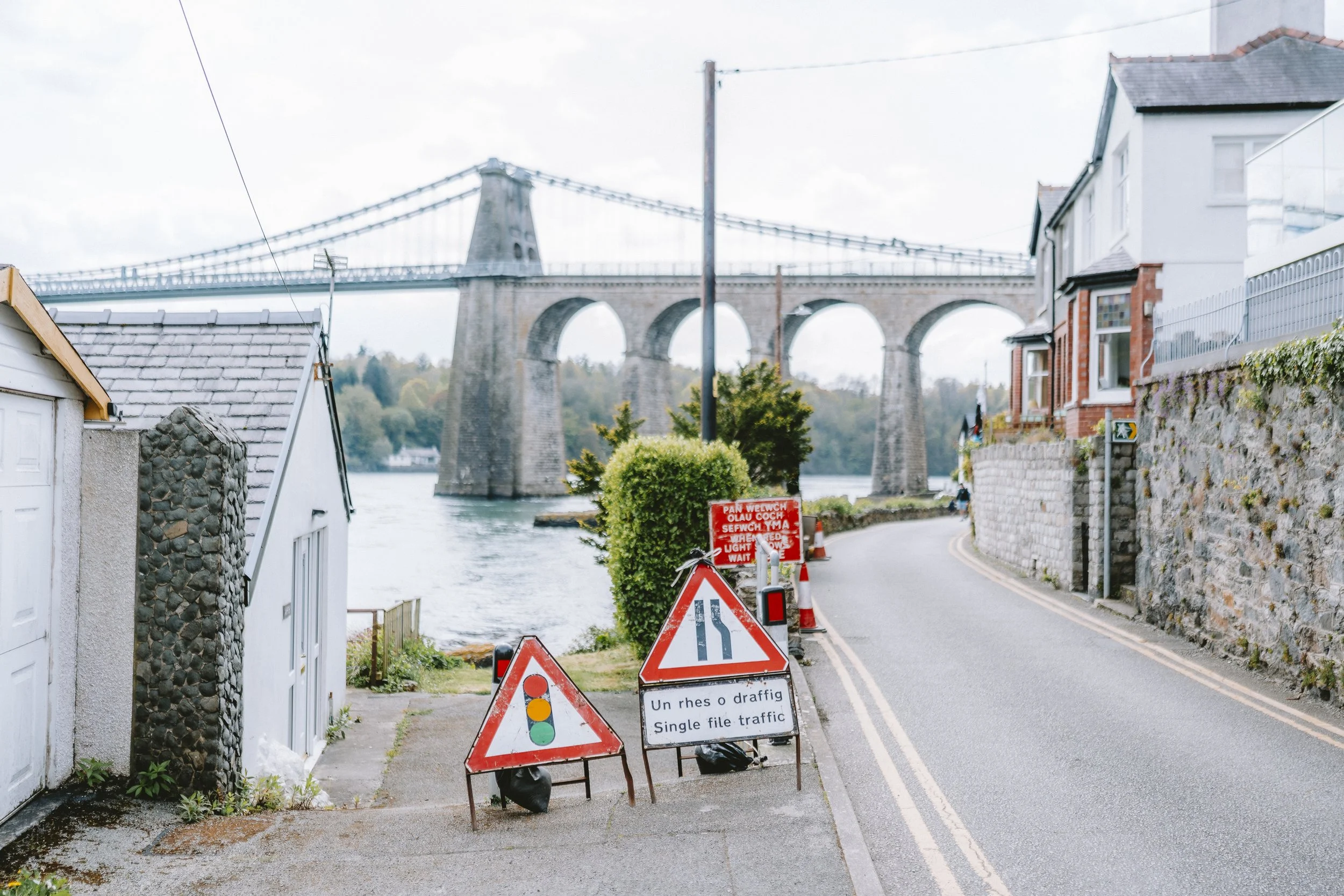 Street with roadwork signs and construction cones in a small town near a river, with a bridge in the background.