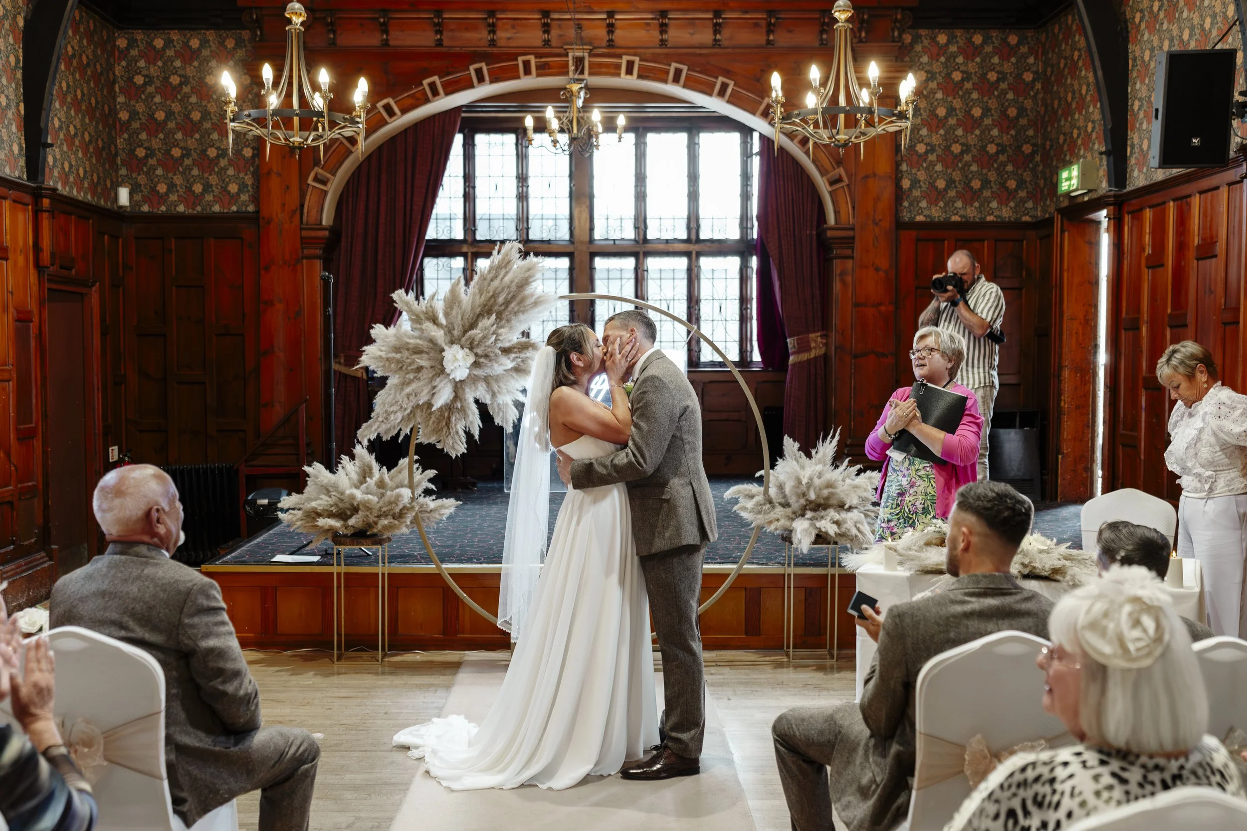 A couple getting married and kissing at their wedding ceremony in a decorated indoor venue, with family and guests watching, and a photographer taking photos.