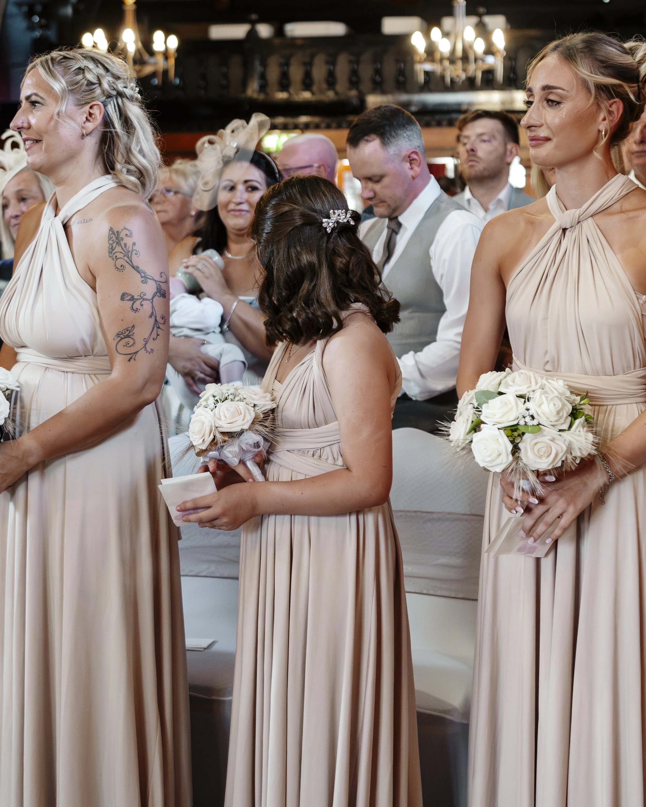 Wedding ceremony with bridesmaids holding white bouquets, women dressed in matching light beige dresses, and a man in a suit among guests seated in a decorated venue with warm lighting.