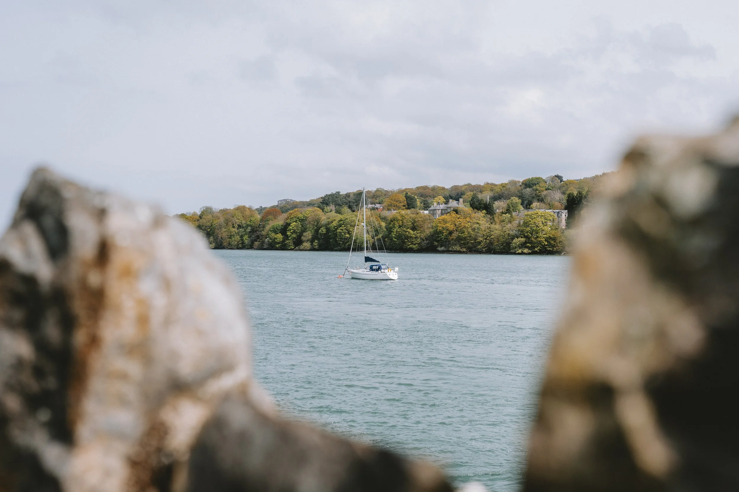 A sailboat floating on a body of water, viewed through rocks or a break in a natural barrier, with a hillside covered in trees and houses in the background under a cloudy sky.