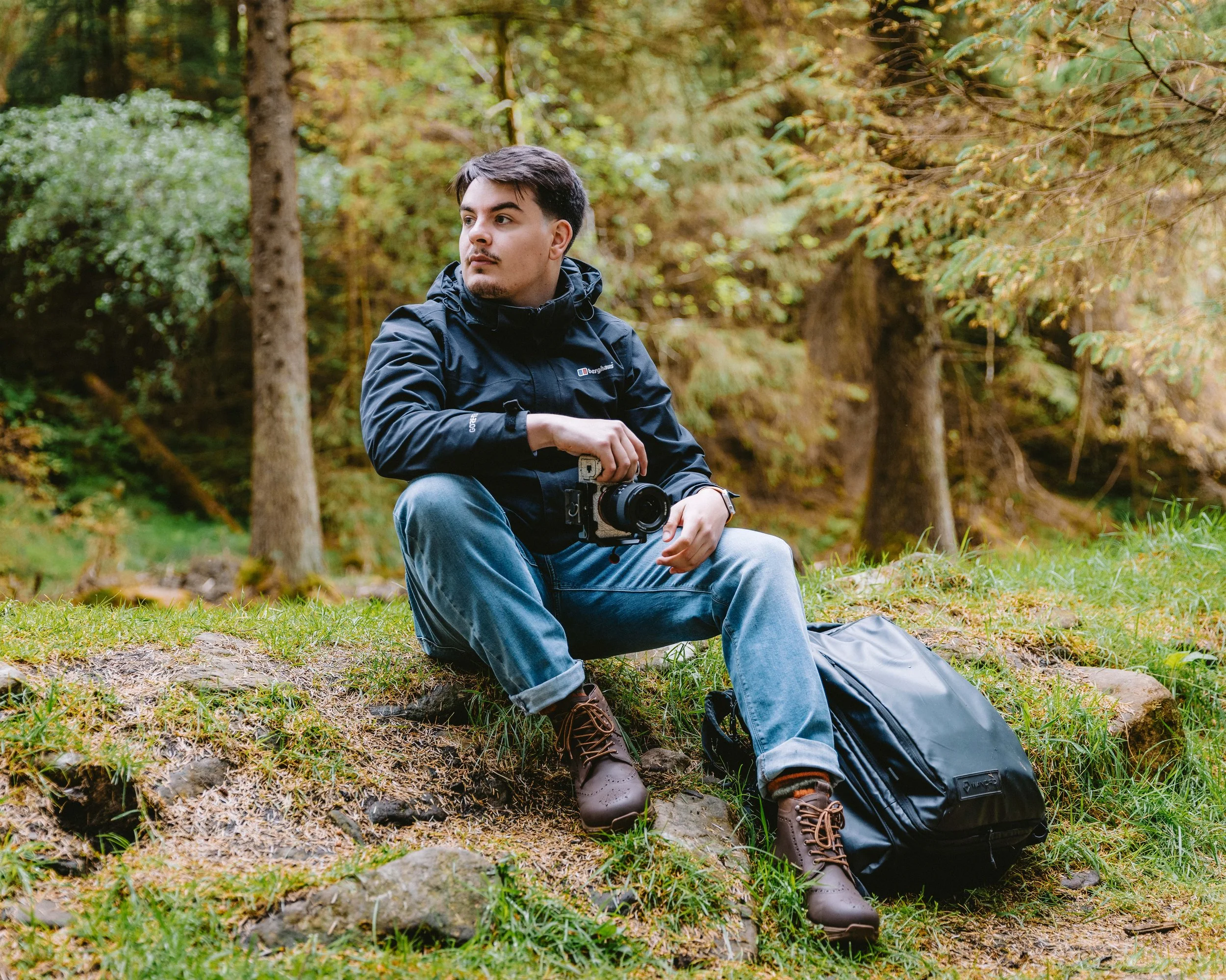 A young man with dark hair, wearing a black jacket and jeans, is sitting outdoors in a forest, holding a camera in his lap. A black camera bag is on the ground beside him. Trees with green and yellow leaves surround him, indicating autumn season.