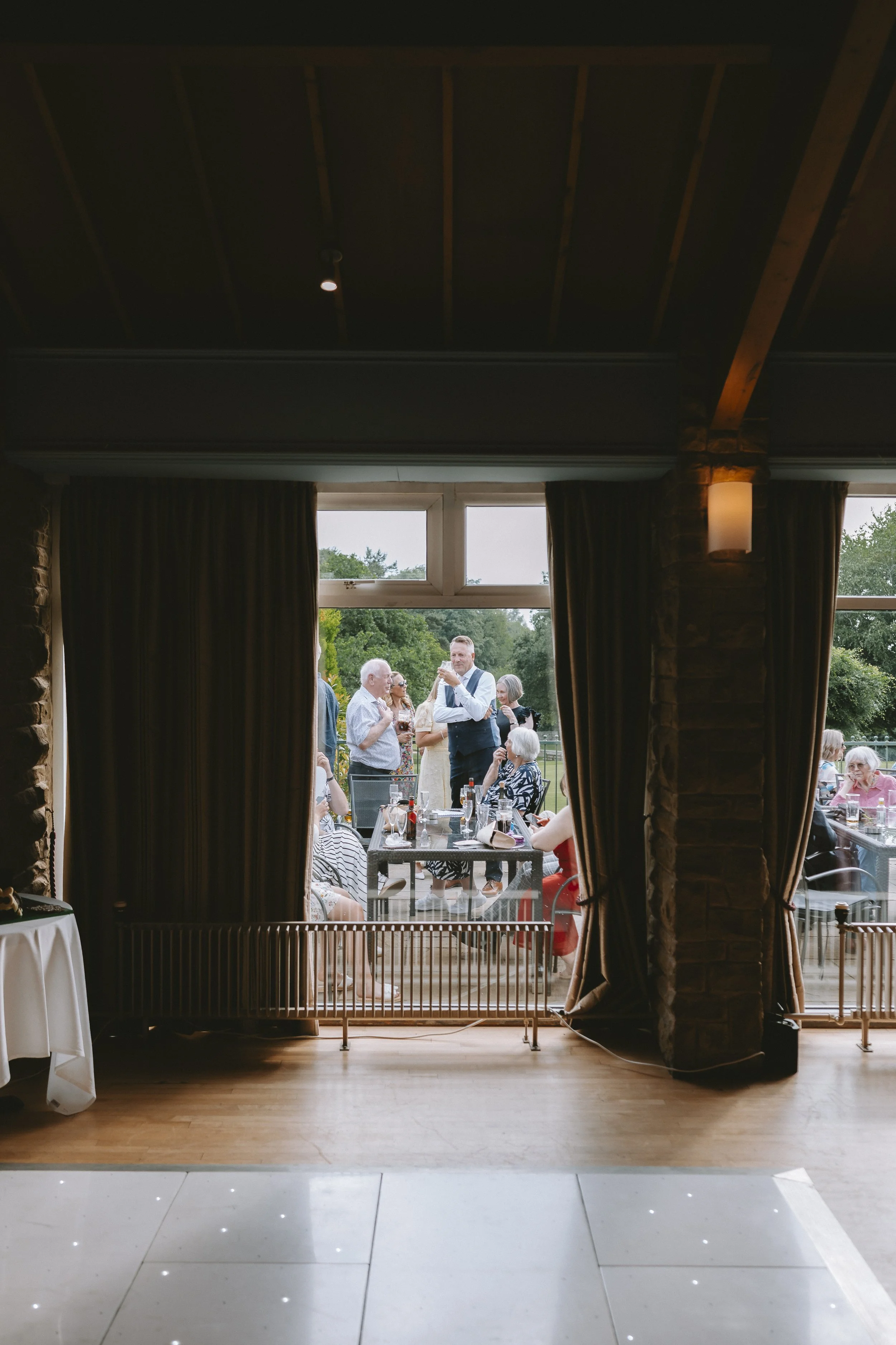 Guests socializing outdoors at a wedding reception, seen through an open window with dark curtains, at a venue with brick walls and wooden ceiling.