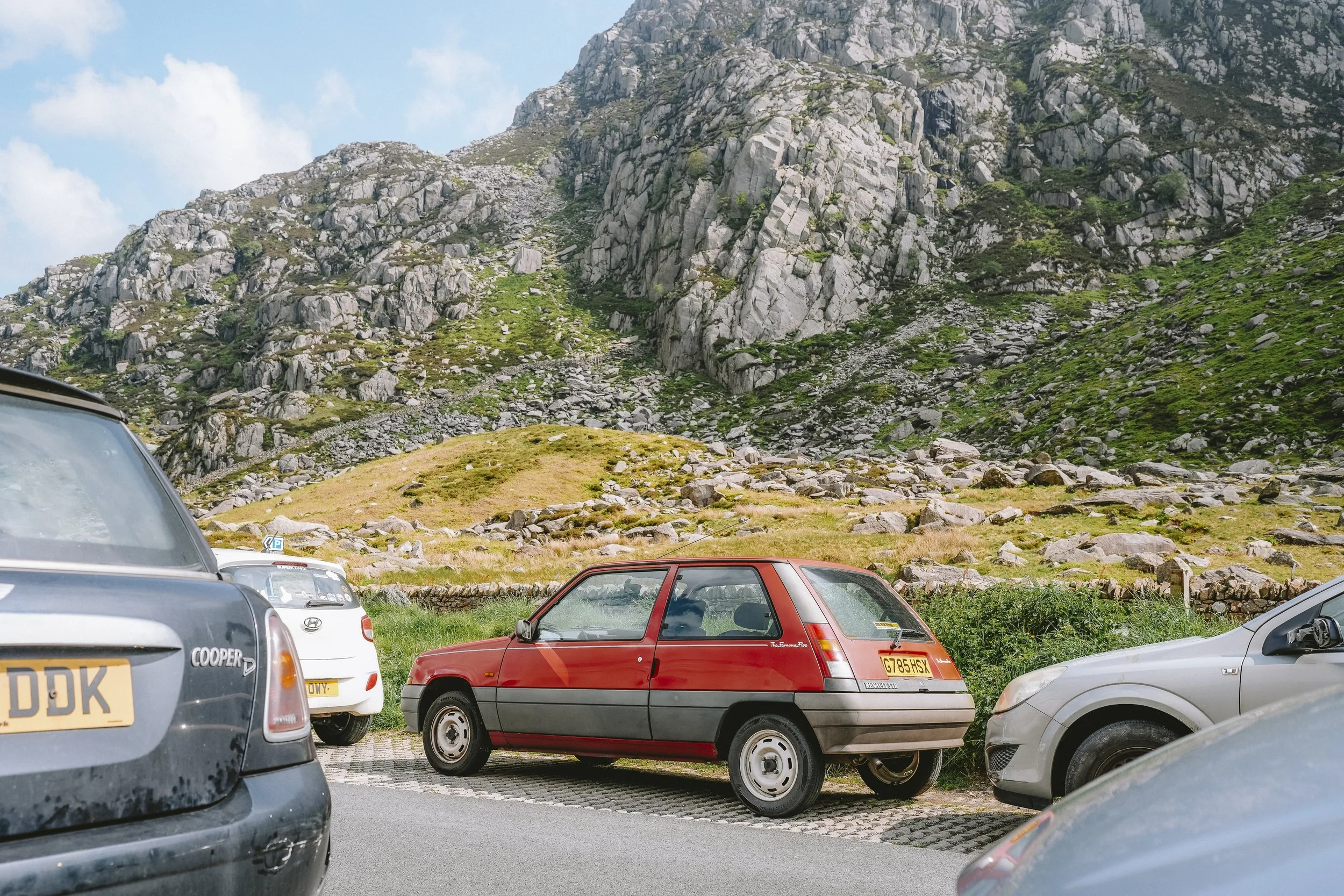 A parking lot with multiple cars parked in front of a mountainous landscape with rugged, rocky hills covered in patches of green vegetation and a blue sky with some clouds.