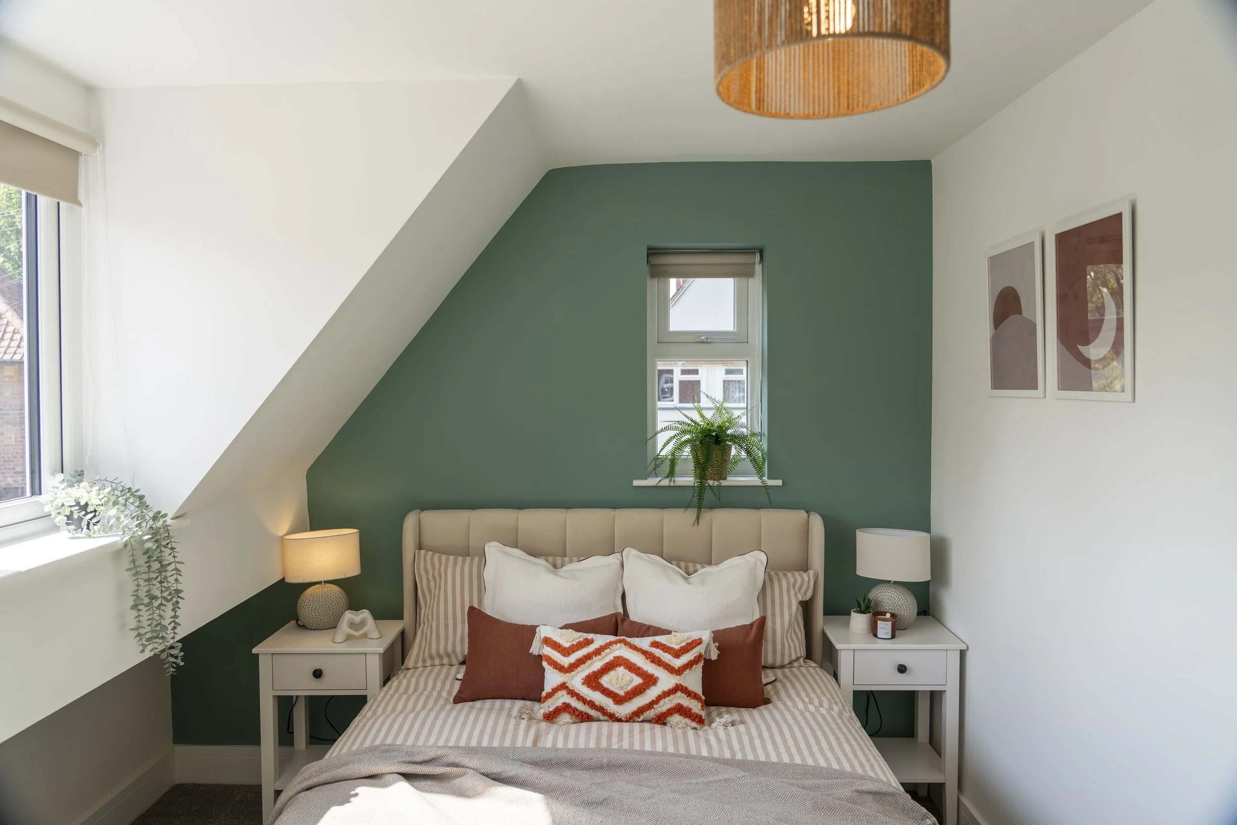 A cozy bedroom with a double bed featuring a beige headboard, striped beige bedding, and white pillows. The bed has a rust-colored pillow and a decorative orange and white pillow. Two white nightstands with lamps flank the bed. The wall behind the be