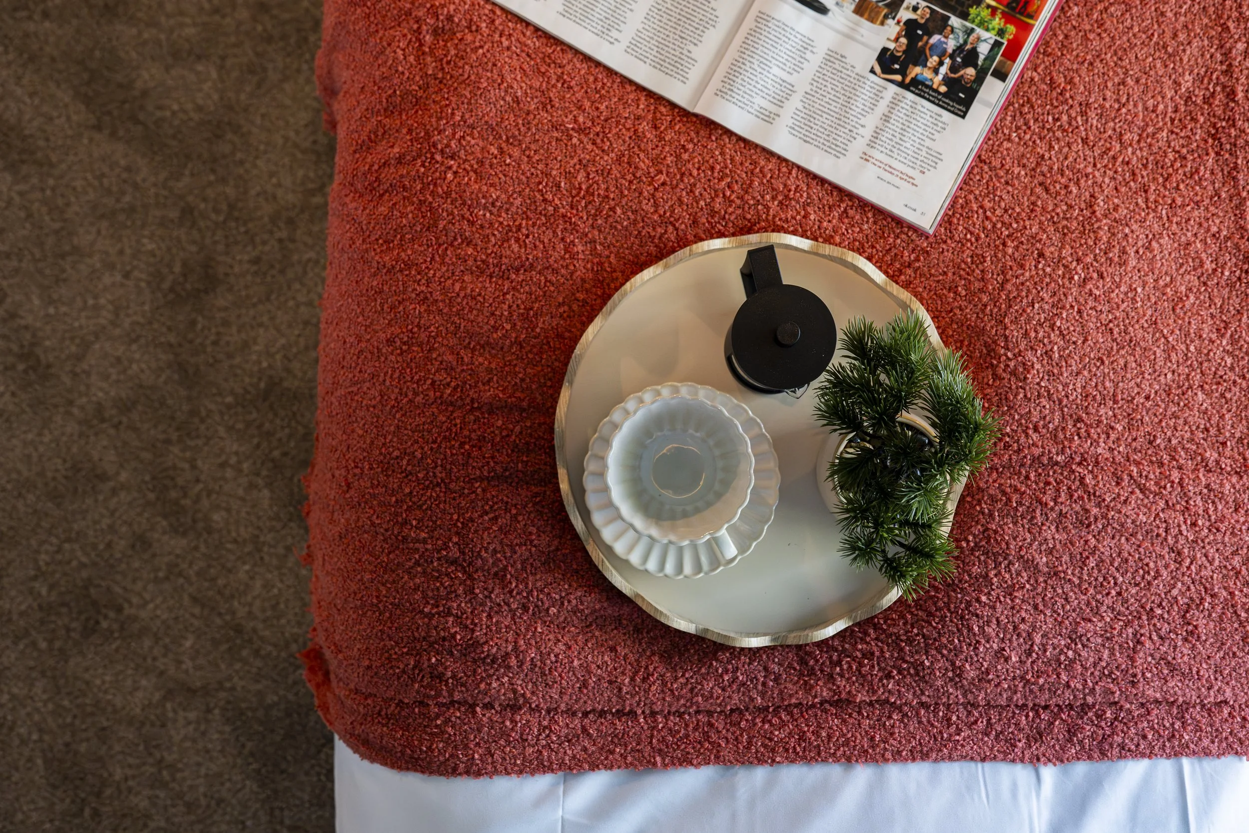 A round serving tray with white scalloped ceramic bowls, a small potted plant, and a black teapot, placed on a textured red tablecloth with an open magazine nearby.
