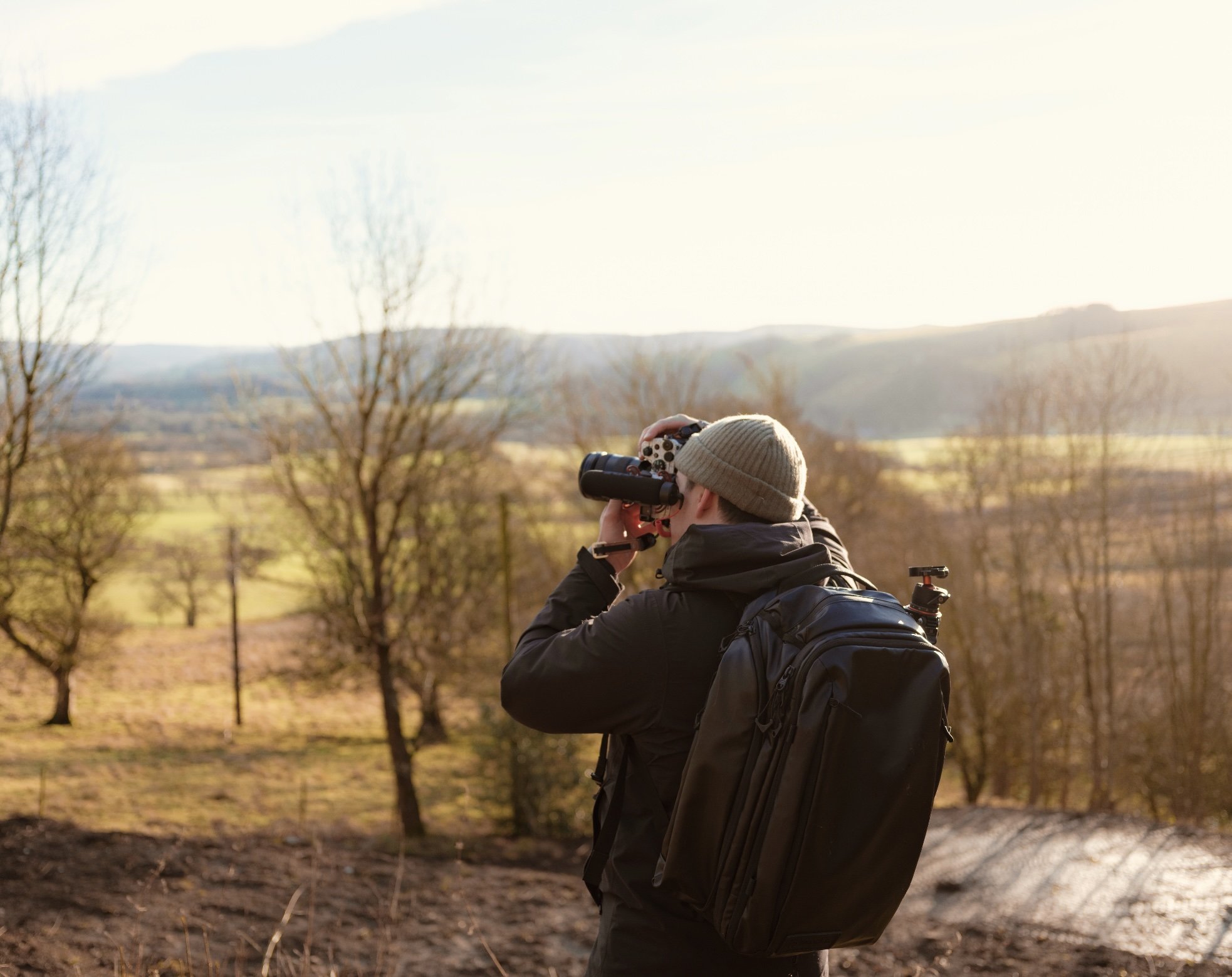 Person wearing a beige beanie hat and dark jacket taking a photo with binoculars outdoors during sunset, with trees and mountains in the background.