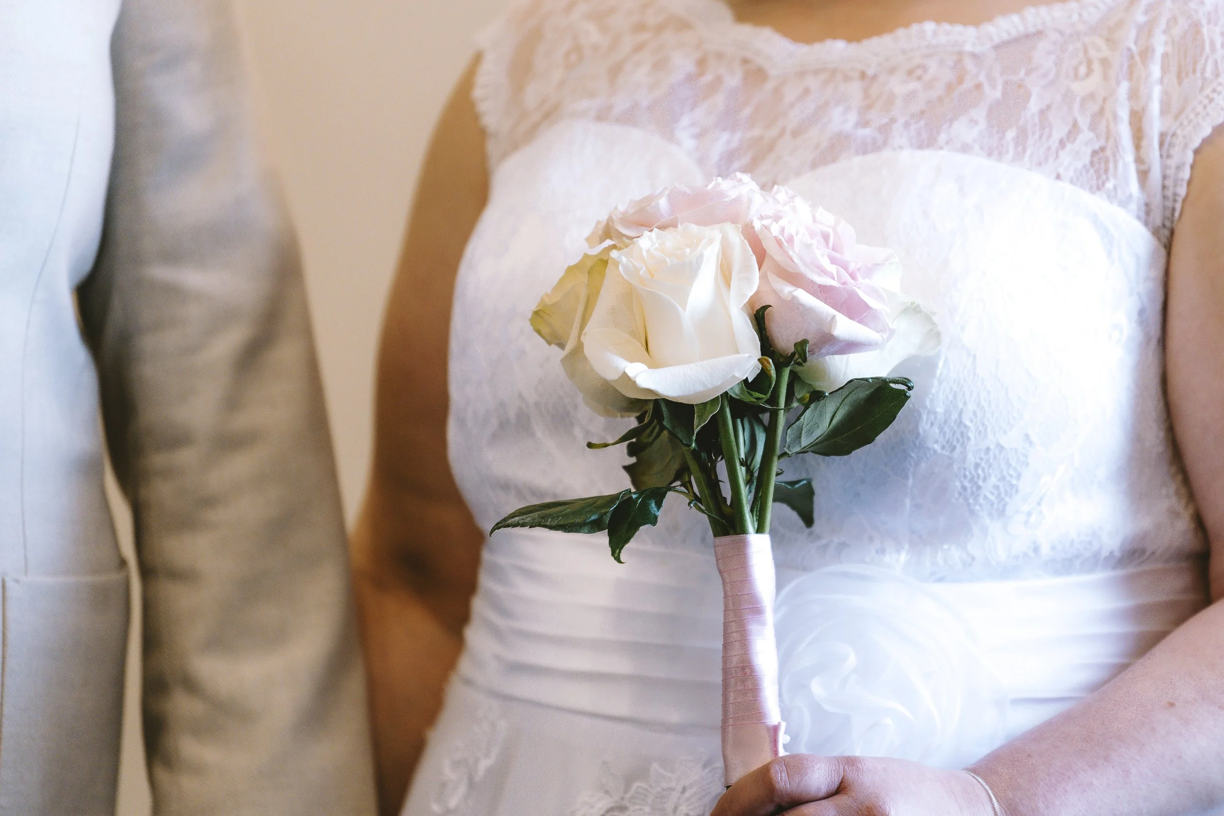 A person dressed in white lace attire holding a bouquet of white and pink roses wrapped in pink ribbon, standing indoors with others nearby.