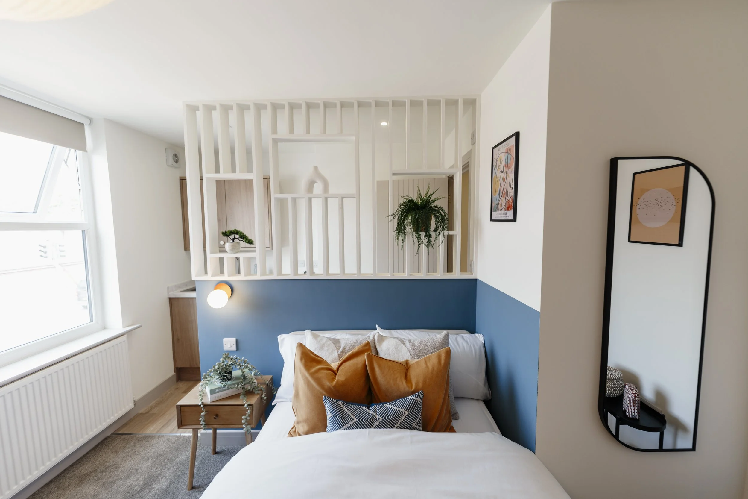 A cozy bedroom with a white bed, brown and black accent pillows, and a small wooden side table with books and a plant. The room features white walls, a blue accent wall, framed artwork, a window with blinds, and decorative shelving with plants and va