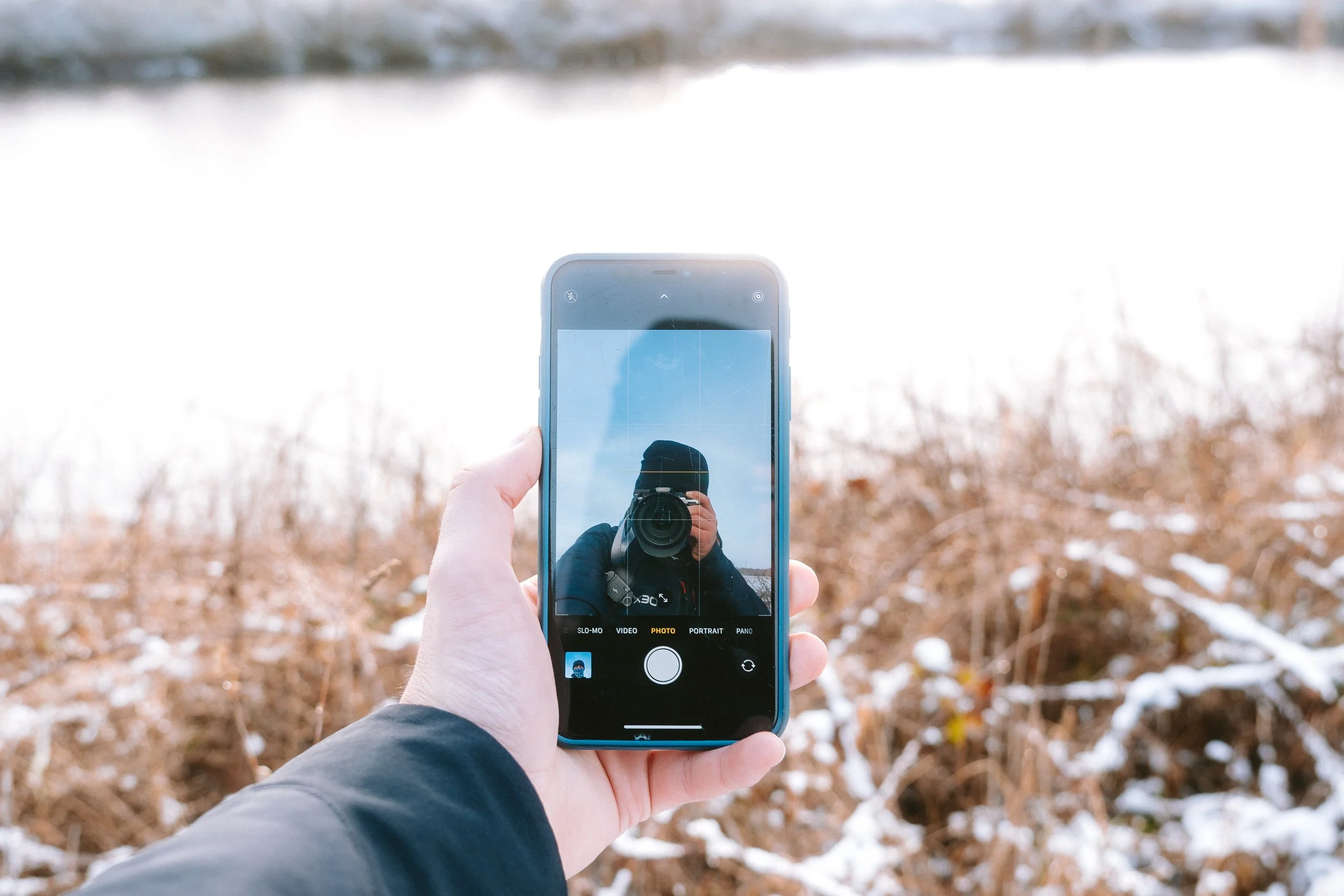 Person holding a smartphone taking a photo in a winter landscape with snow and brown shrubs.