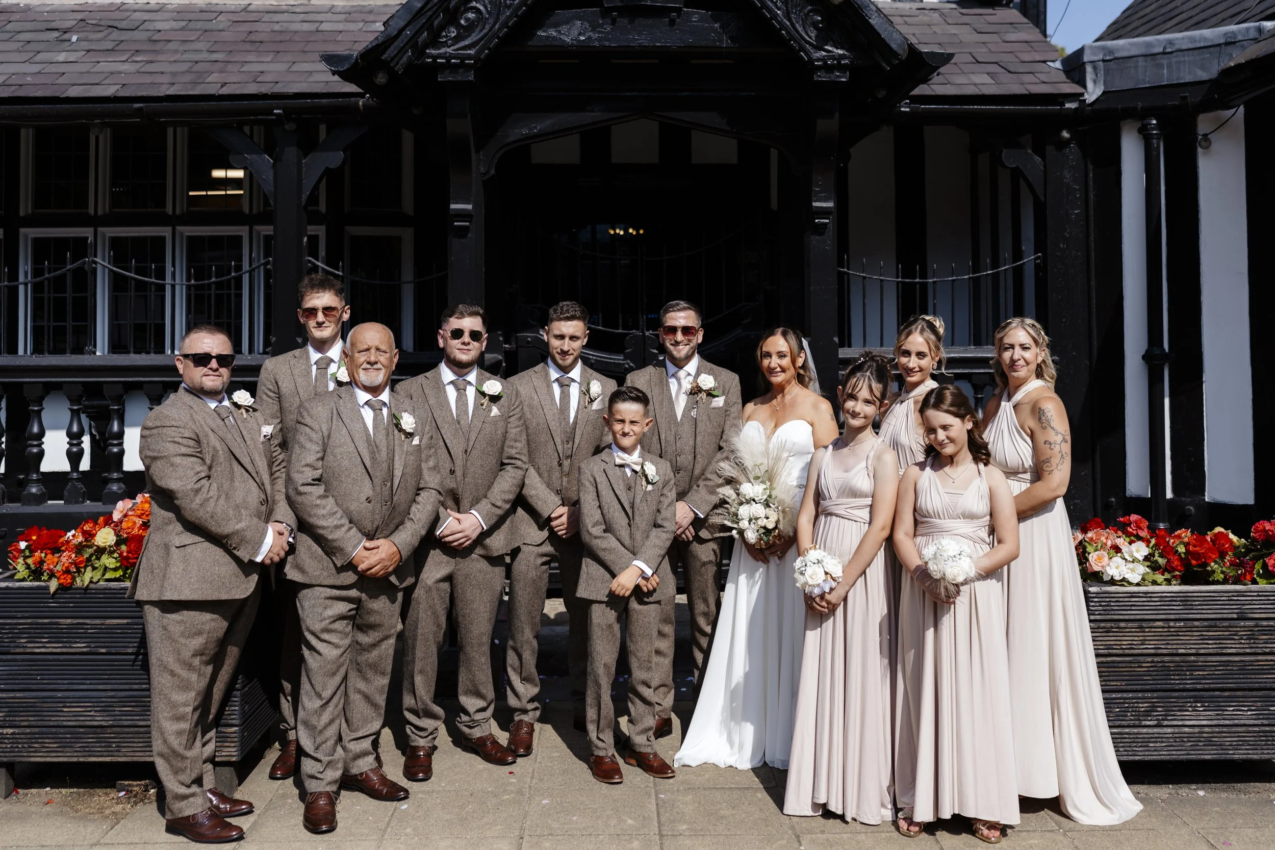 Group of bridesmaids, groomsmen, bride, and one young boy posing together outdoors for a wedding photo in front of a black building with flower arrangements.