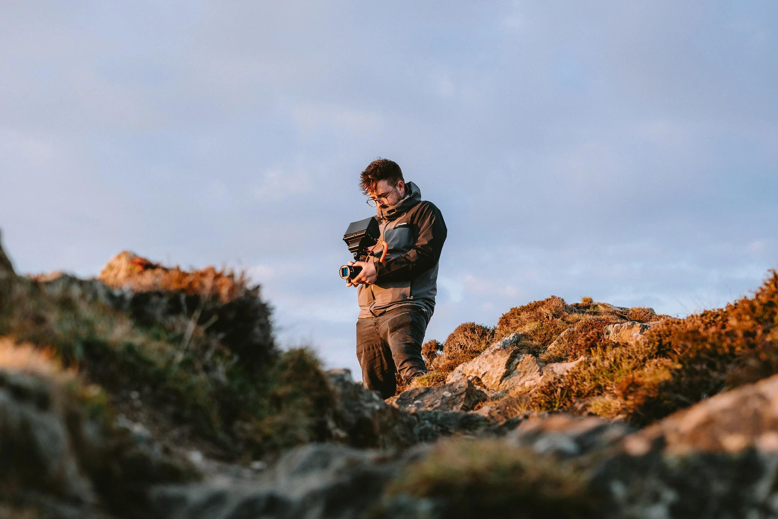 A man wearing glasses and outdoor clothing stands on rocky terrain, looking down at a device in his hands, with a cloudy sky in the background.