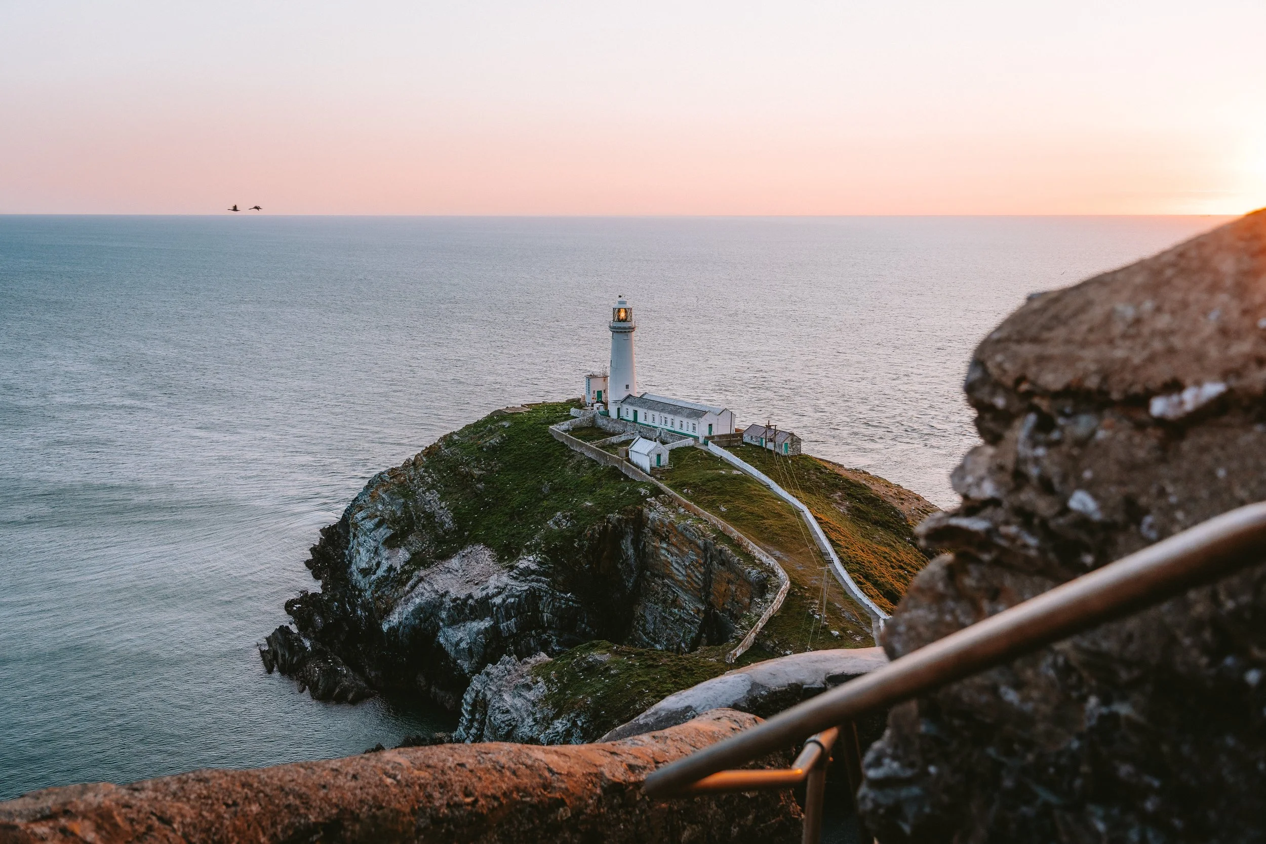 A lighthouse on a small rocky island surrounded by water, with a pink and orange sunset sky and two birds flying in the distance.