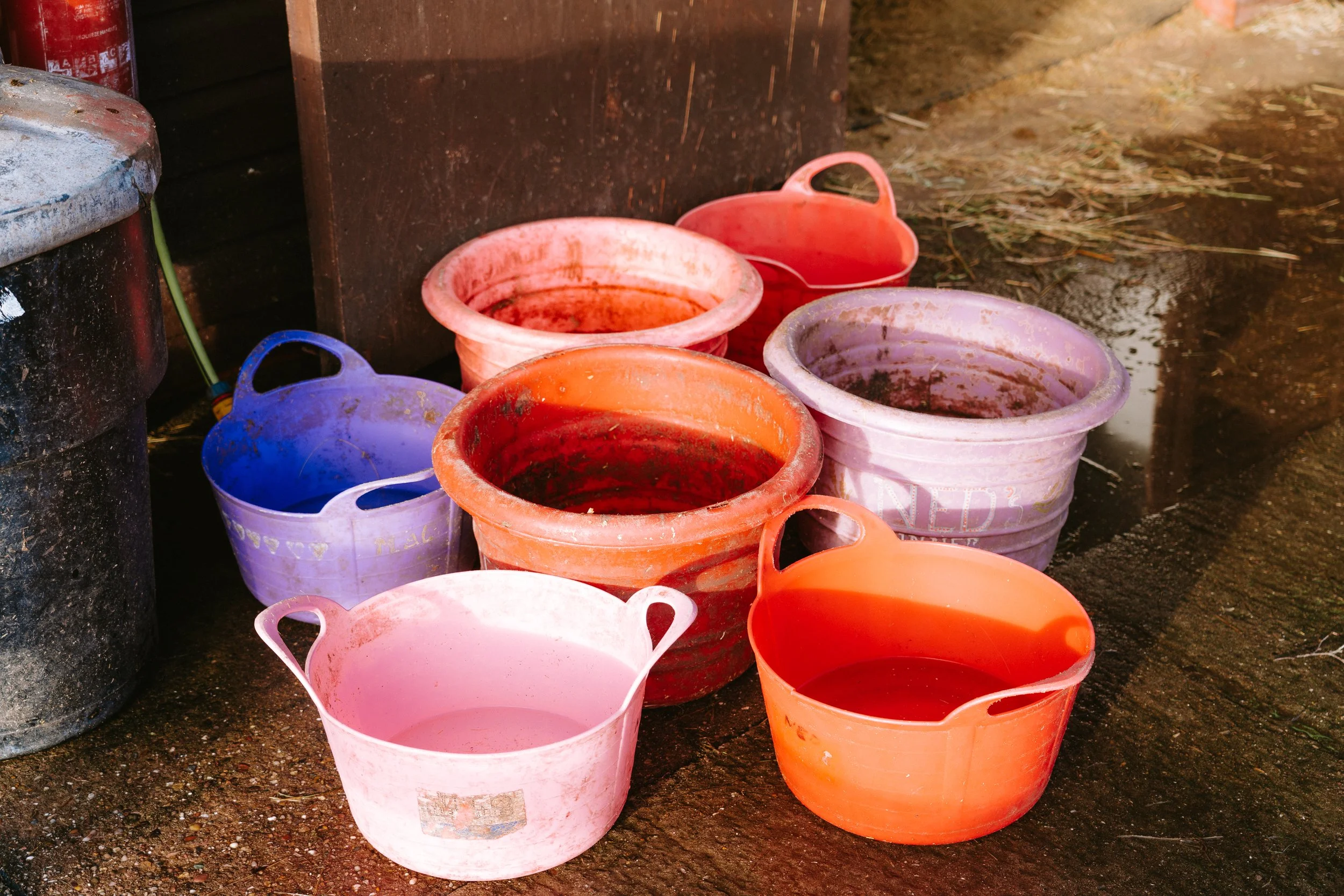 Colorful plastic buckets, mostly pink and orange, stacked and dirty, on a muddy ground beside a black container and a large black object.