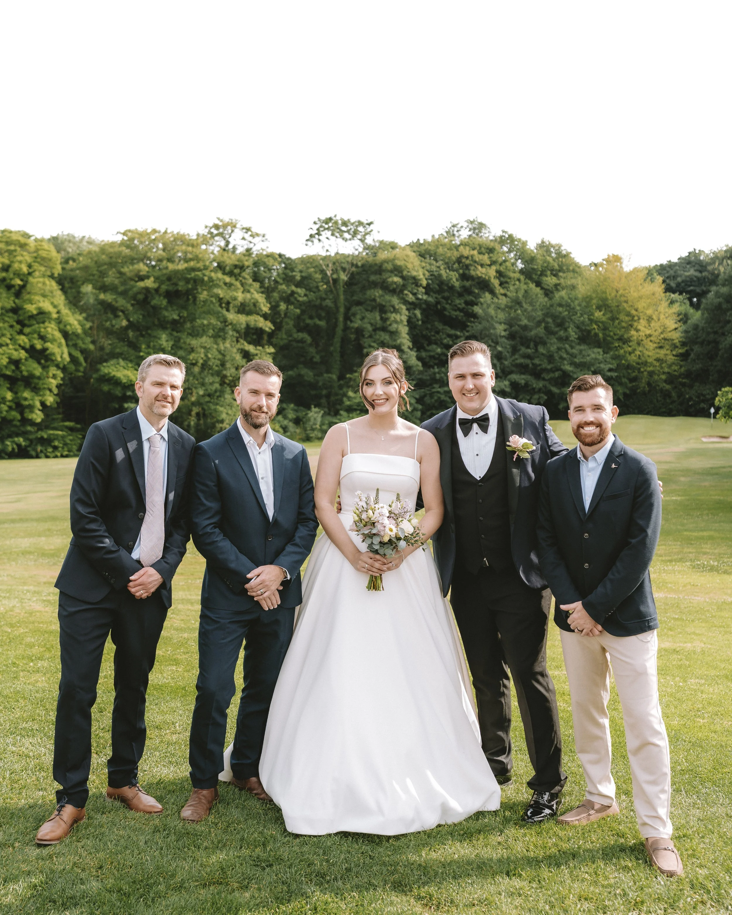 Group of five people dressed in formal attire, standing on a grassy area with trees in the background, possibly at a wedding or special event.