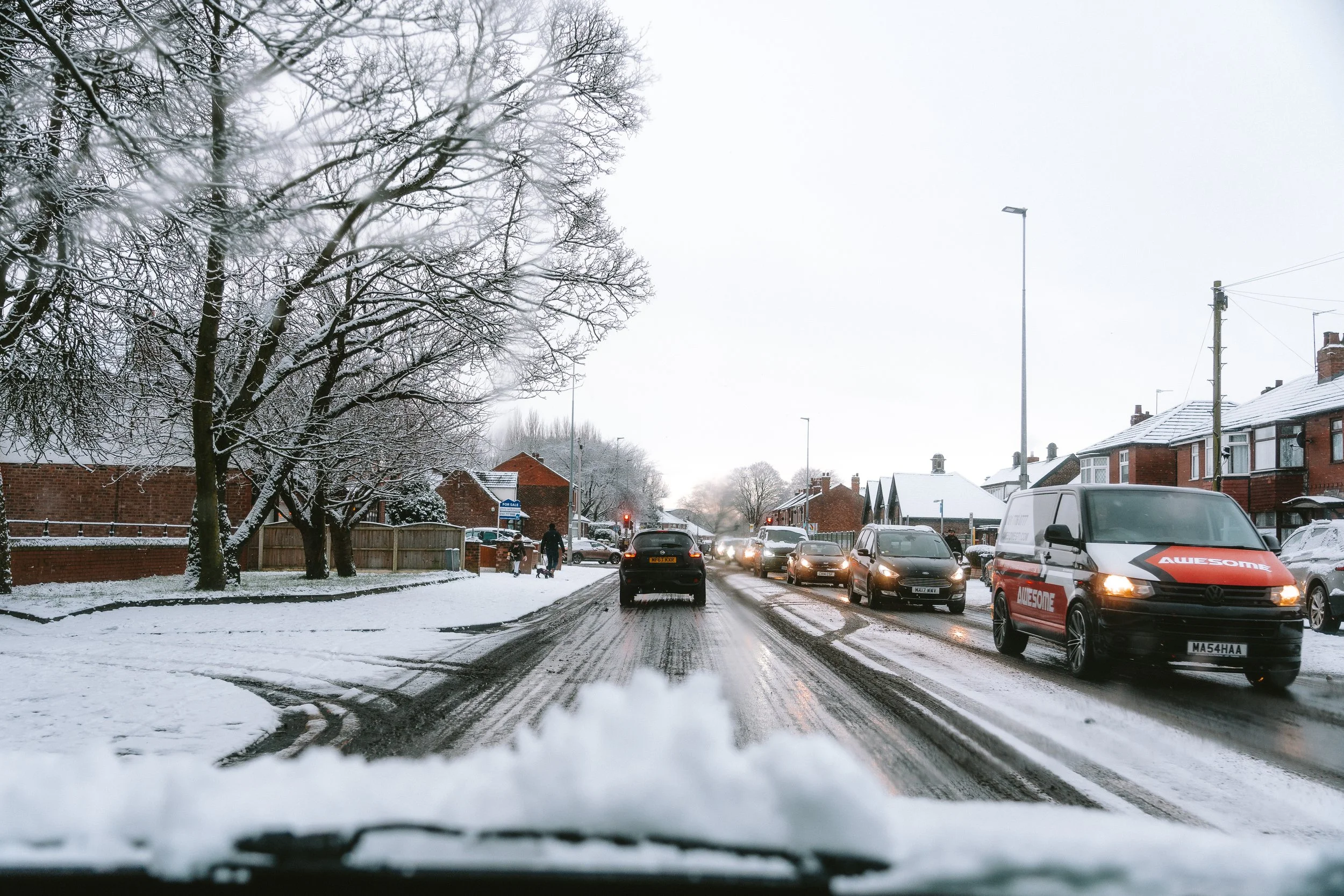 A snowy street scene with cars driving and parked, people walking, and houses with snow-covered roofs, under an overcast sky.