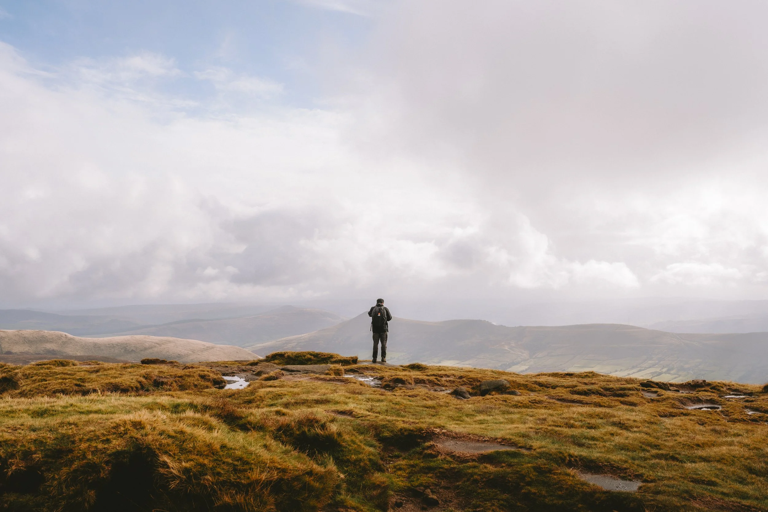 A person with a backpack standing on a grassy hilltop looking at distant mountains and a cloudy sky.