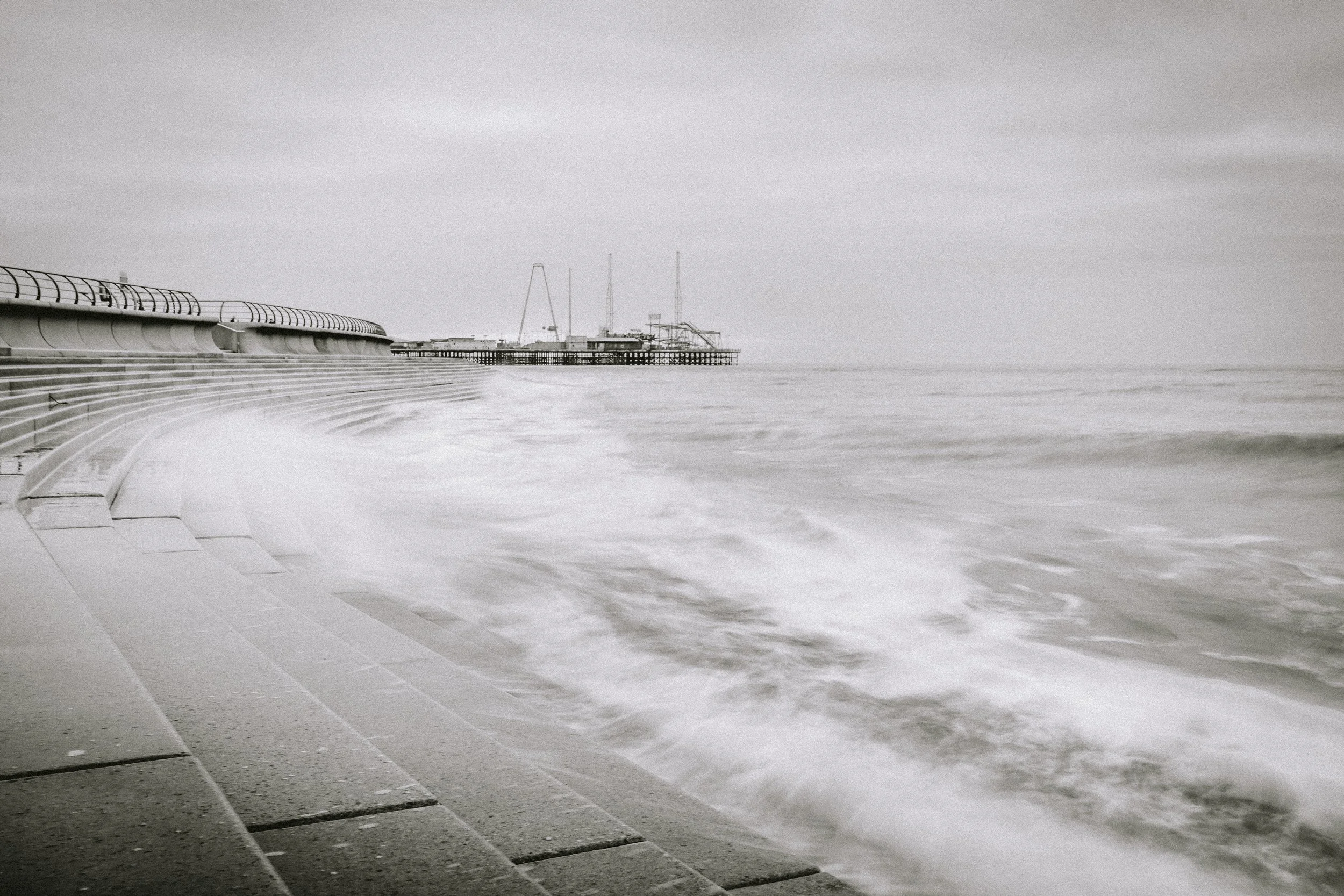 A cloudy sky over a beach with ocean waves crashing against a concrete seawall and a pier with structures in the distance.