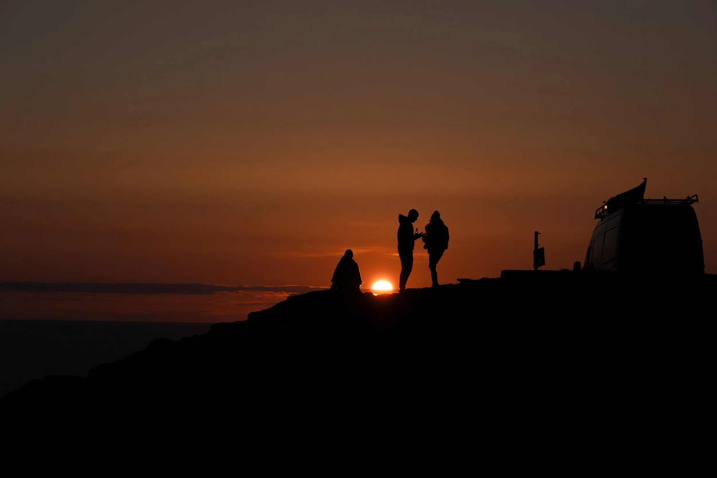 Silhouettes of three people standing on a cliff near the ocean during sunset, with a vehicle parked nearby and the sun on the horizon.