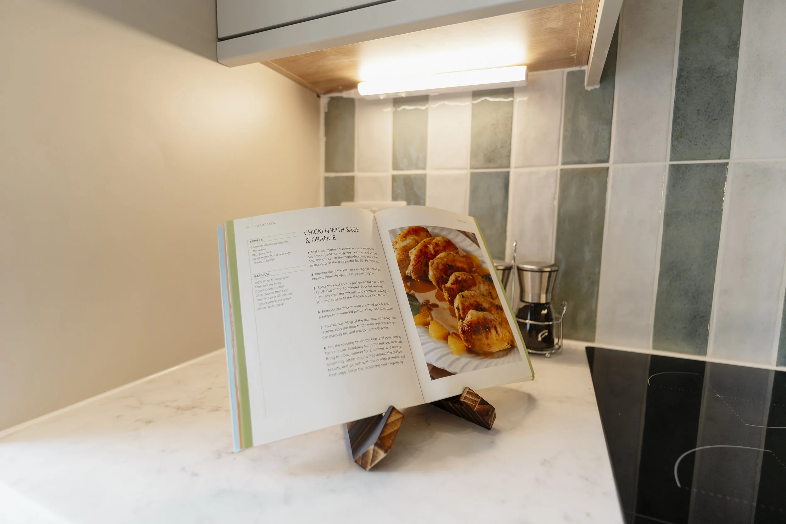 Open cookbook displaying a chicken recipe, placed on a kitchen counter with tiled backsplash and a bottle of oil or vinegar in the background.