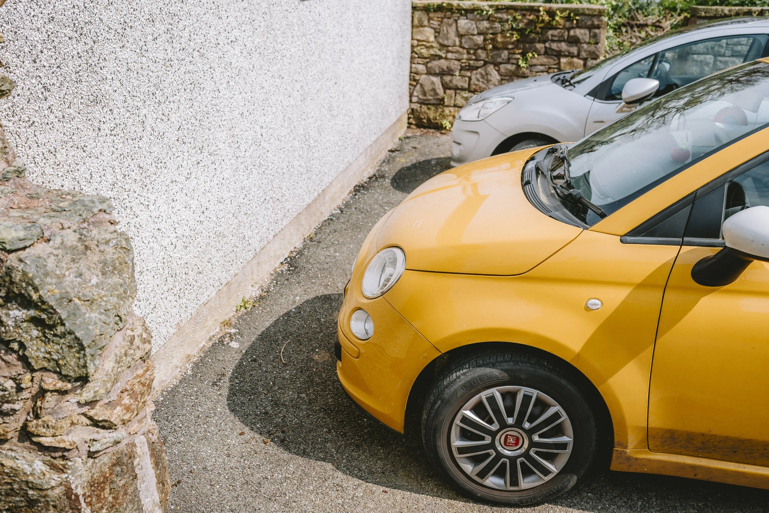 Close-up of the front of a yellow Fiat car parked next to a white car in a parking lot beside a textured stone wall.