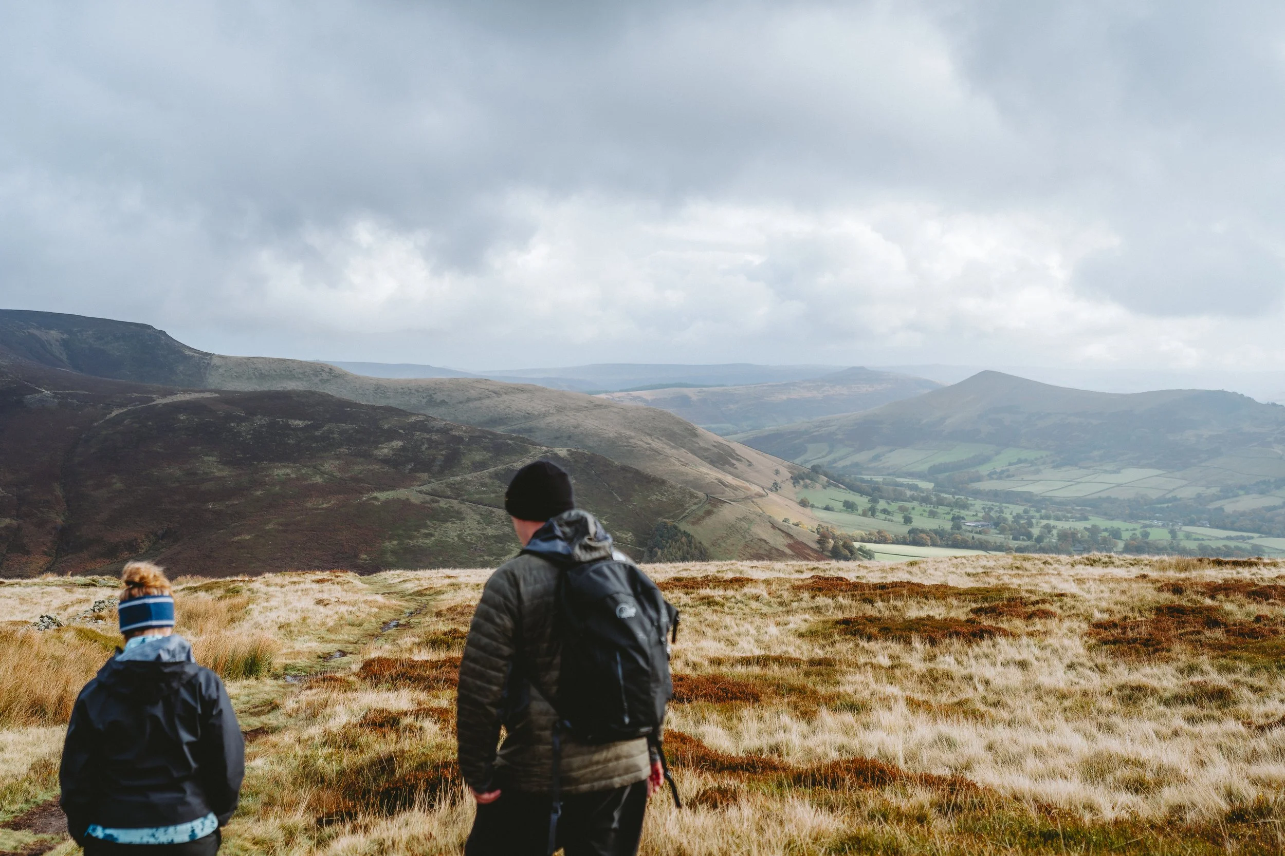 Two children and an adult hiking through a hilly landscape with green fields, trees, and mountainous terrain in the distance under cloudy skies.