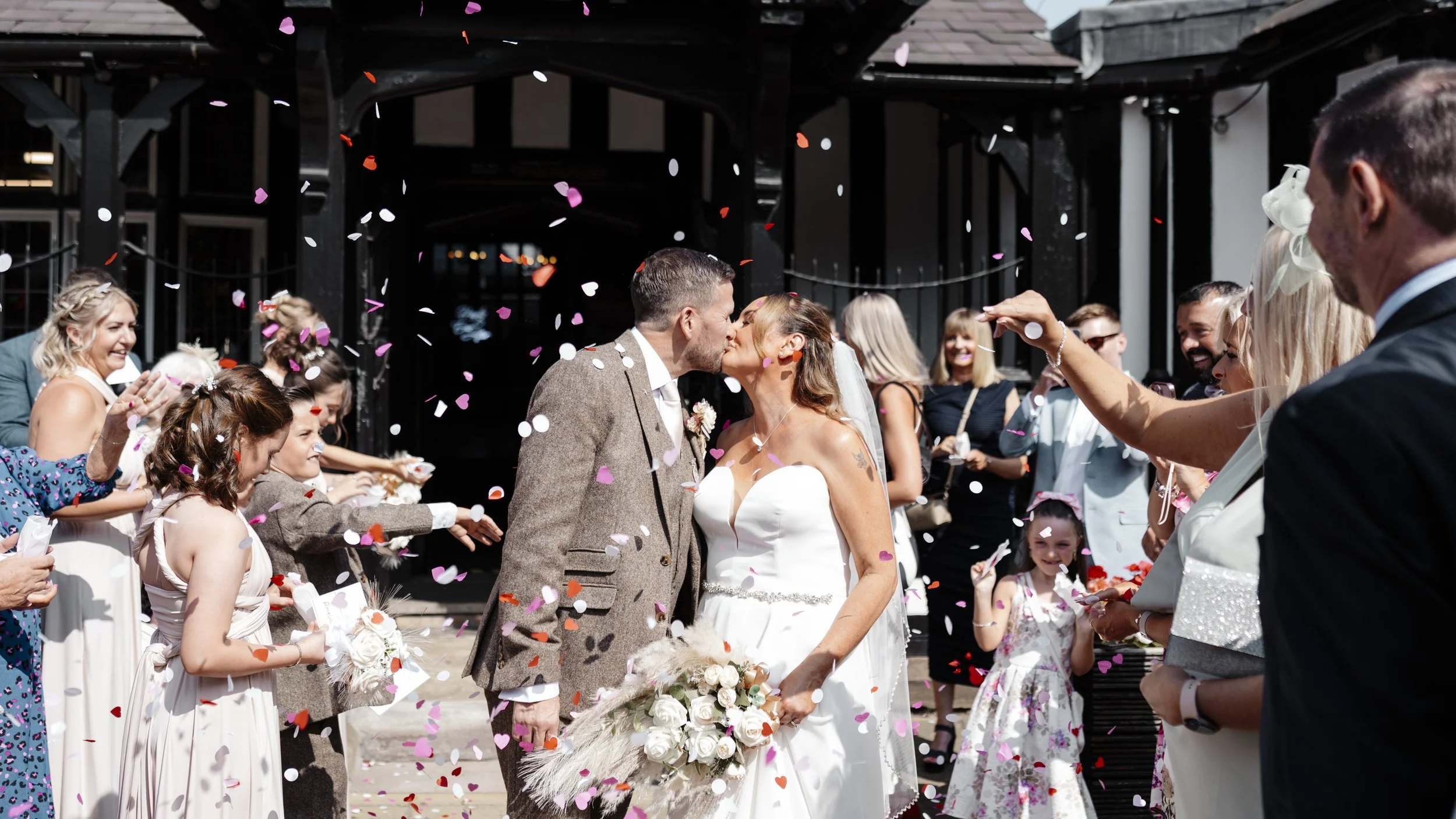 A newlywed couple kisses under falling colorful confetti surrounded by friends and family celebrating at their wedding ceremony.