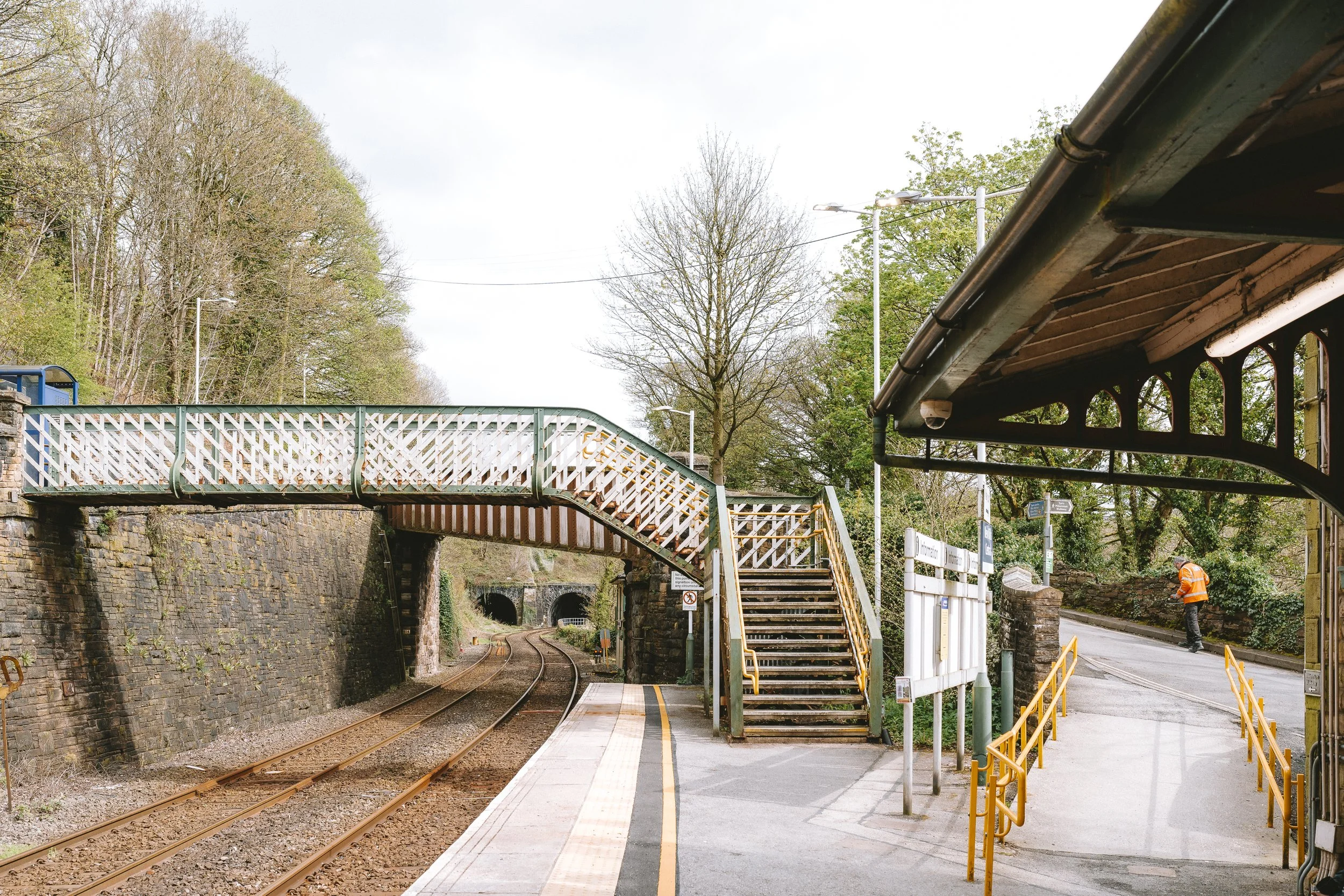 View of a train station platform with stairs leading to an overpass, trees in the background, train tracks underneath, and a worker in orange safety clothing.