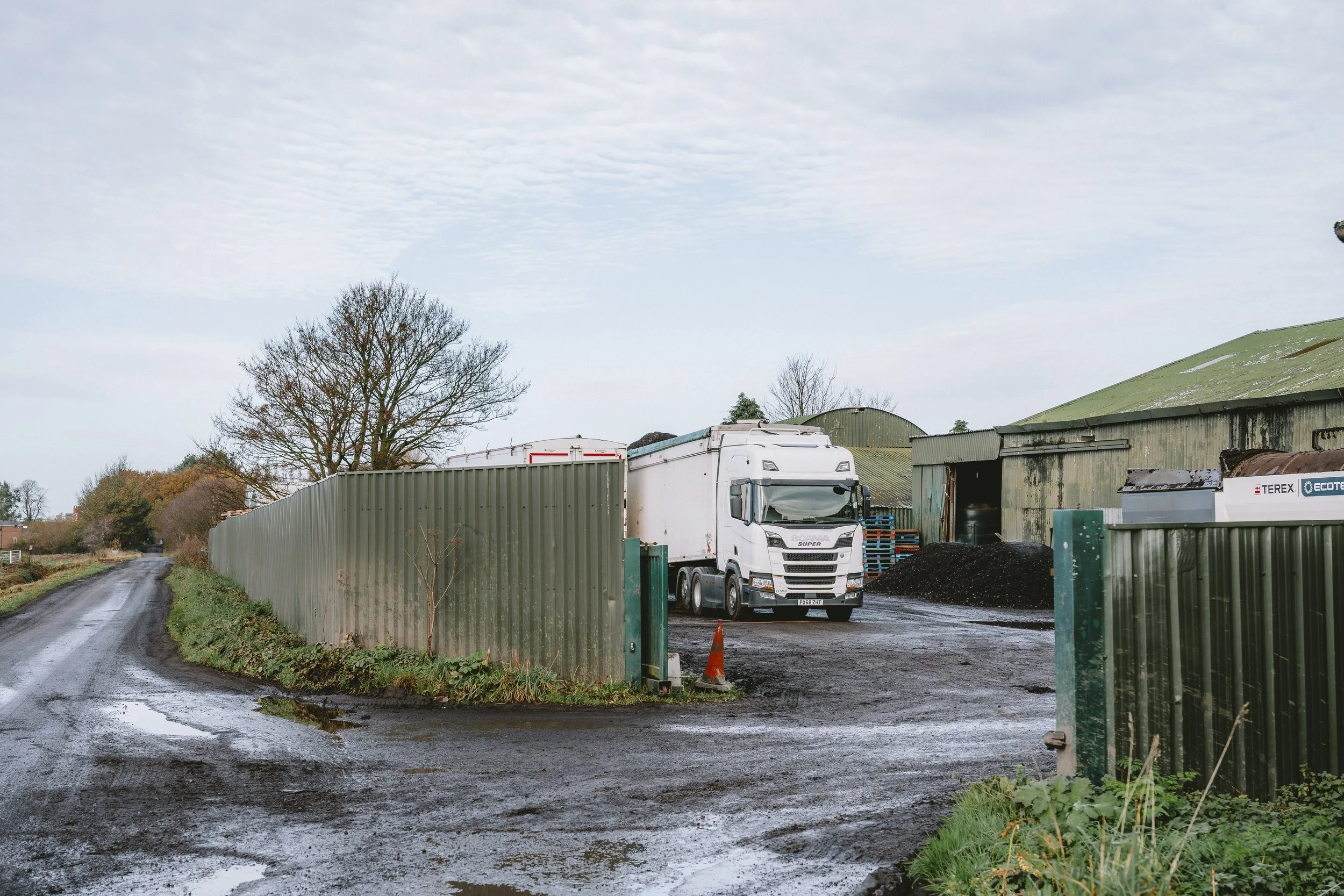 A farm or industrial yard with a white truck parked inside a green metal fence and a large green building with a curved roof in the background. The ground is muddy and wet, with a muddy road leading past the fence.