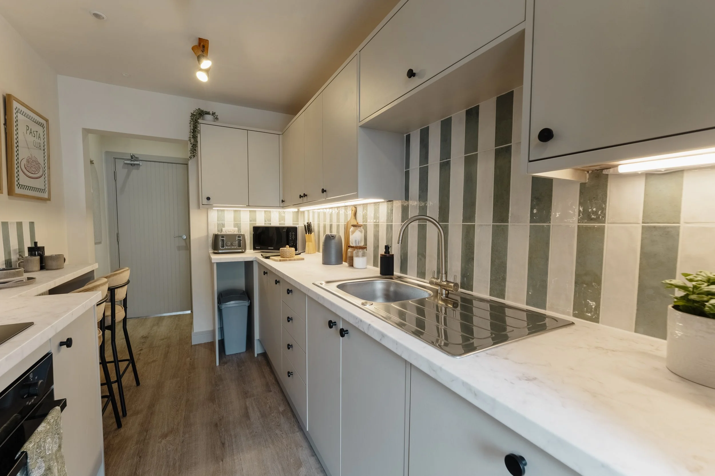 A modern kitchen with white cabinetry, a marble countertop, and a striped green and white tile backsplash. There is a double sink with a stainless steel faucet, small appliances like a microwave and toaster, and decorative items including jars and a 